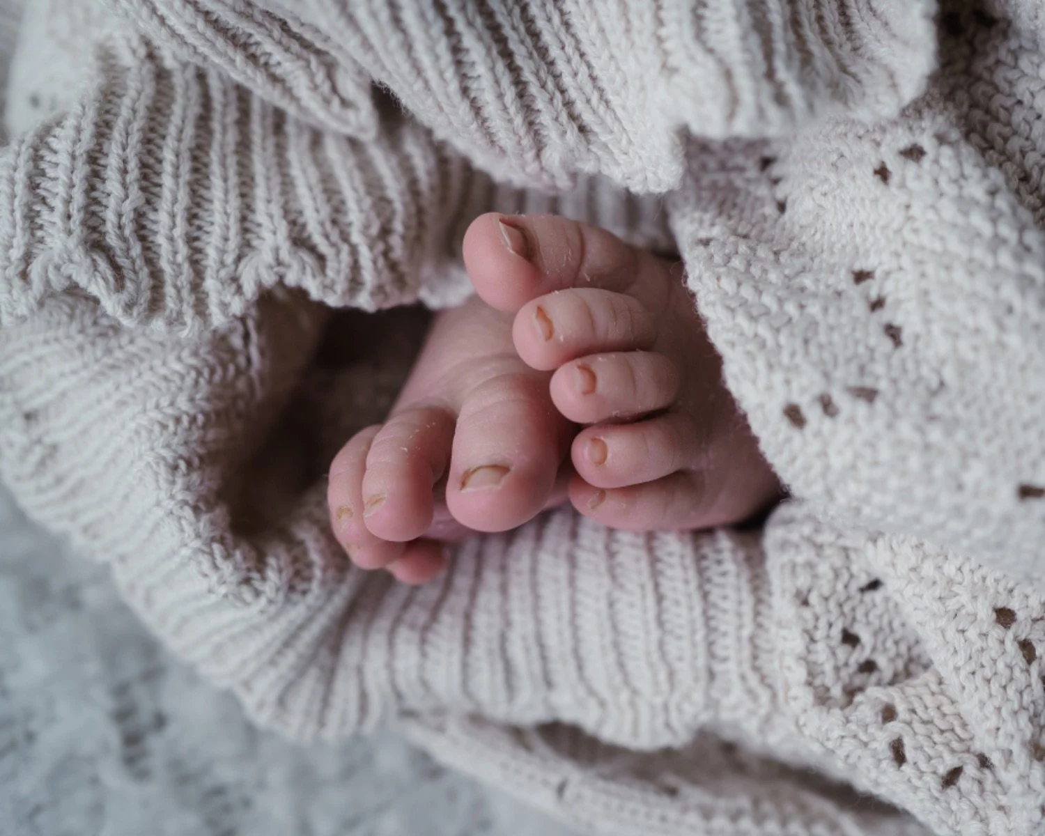Close-up of a newborn baby's tiny hand clutching an adult's finger, wrapped in soft, knitted beige fabric.