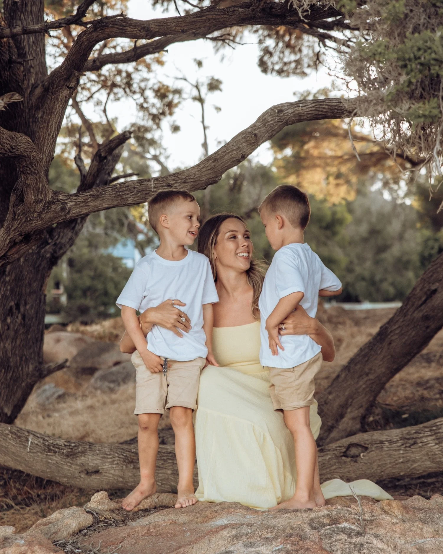 A woman sitting outdoors on a rock, smiling and holding two young boys, all dressed in light-colored clothing, surrounded by trees and natural scenery during sunset.