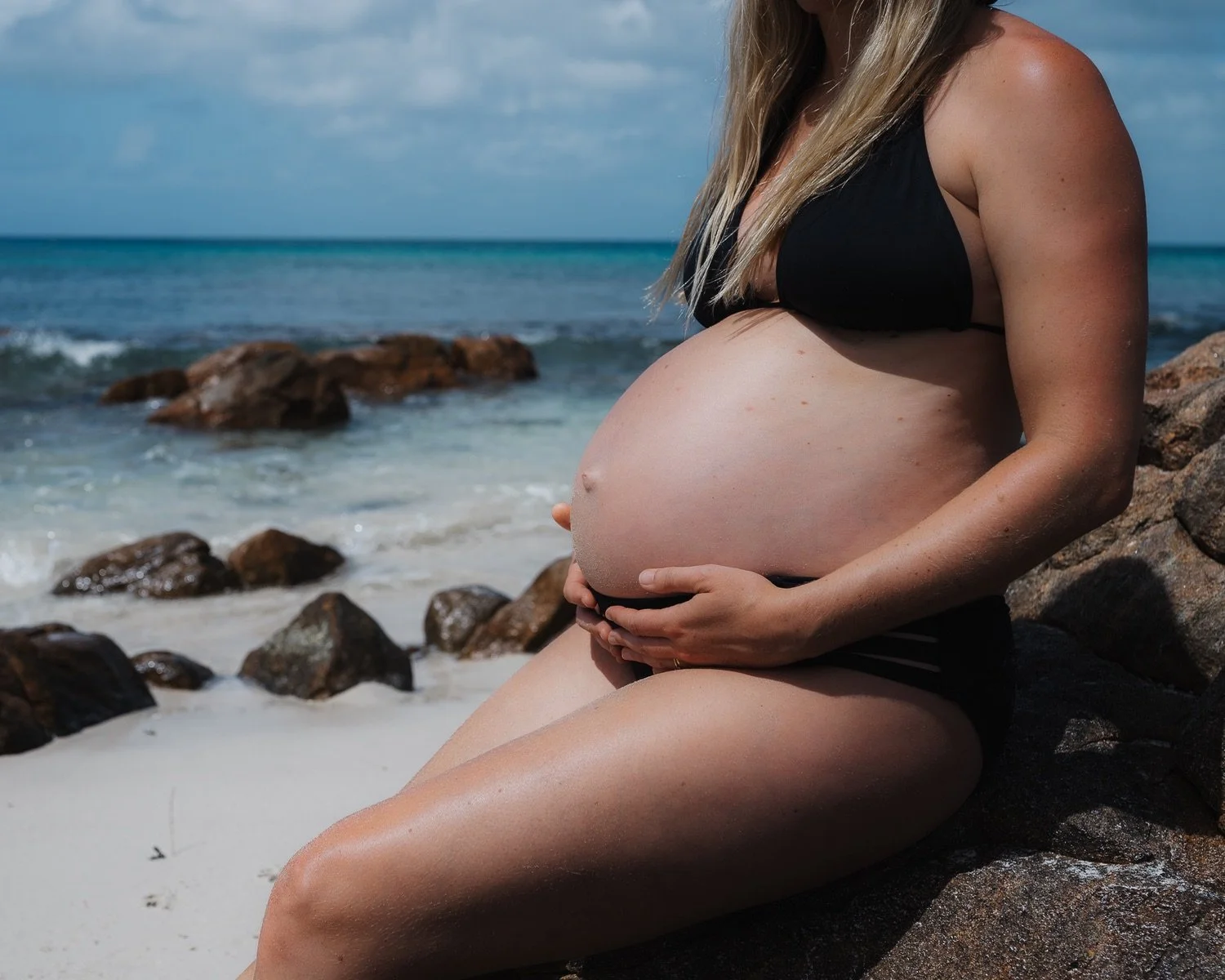 Pregnant woman in a black bikini sitting on rocks at the beach with ocean waves and rocks in the background.