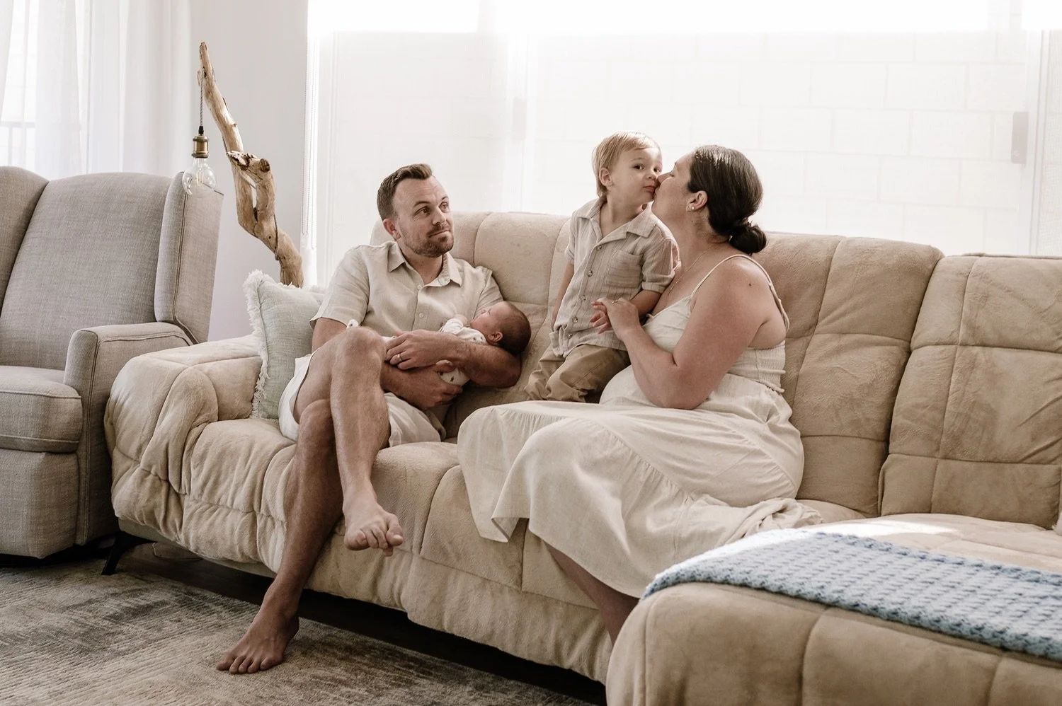 Family sitting on a beige sofa in a bright living room, with a father holding a newborn, a young boy giving a kiss to a woman, all dressed in light-colored casual clothes.