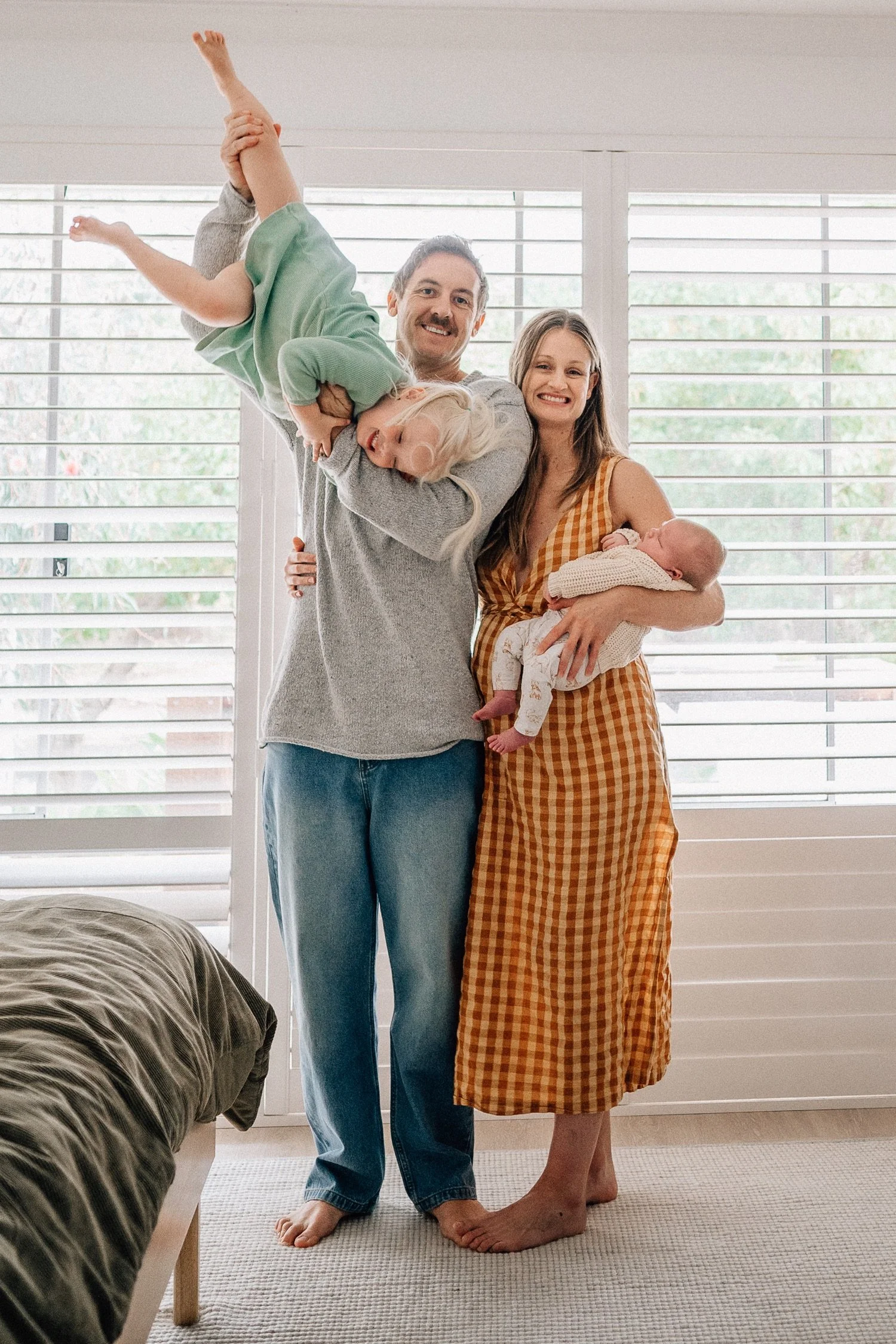 A family of four with two parents, a young girl, and a baby, standing indoors in front of large windows with plantation shutters, smiling and embracing each other. The father is holding the girl upside down, and the mother is holding the baby.