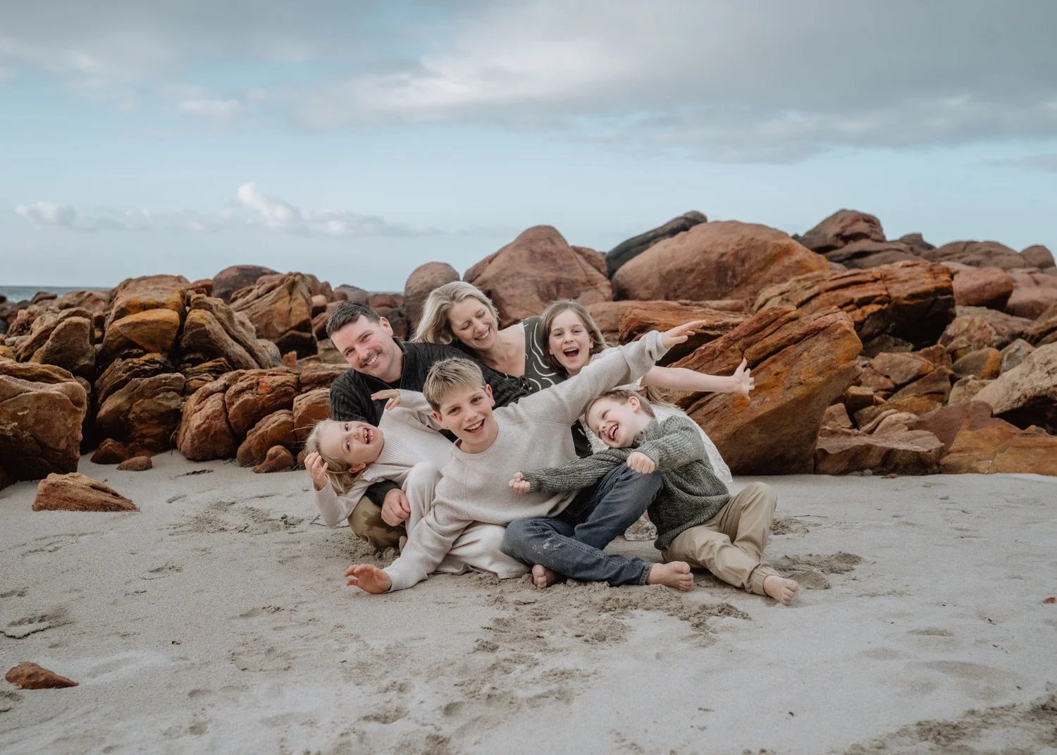 Family of six playing and laughing on a sandy beach with large rocks and cloudy sky in the background.