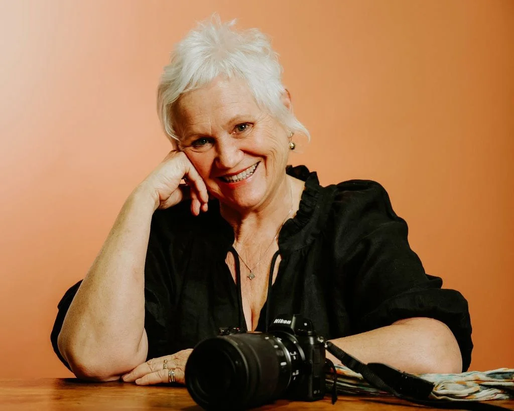 Jill Rogers, of Jill Rogers Photography, with short white hair, sitting at a table with a Nikon camera and some papers, smiling warmly and resting her head on her hand against a gradient background.