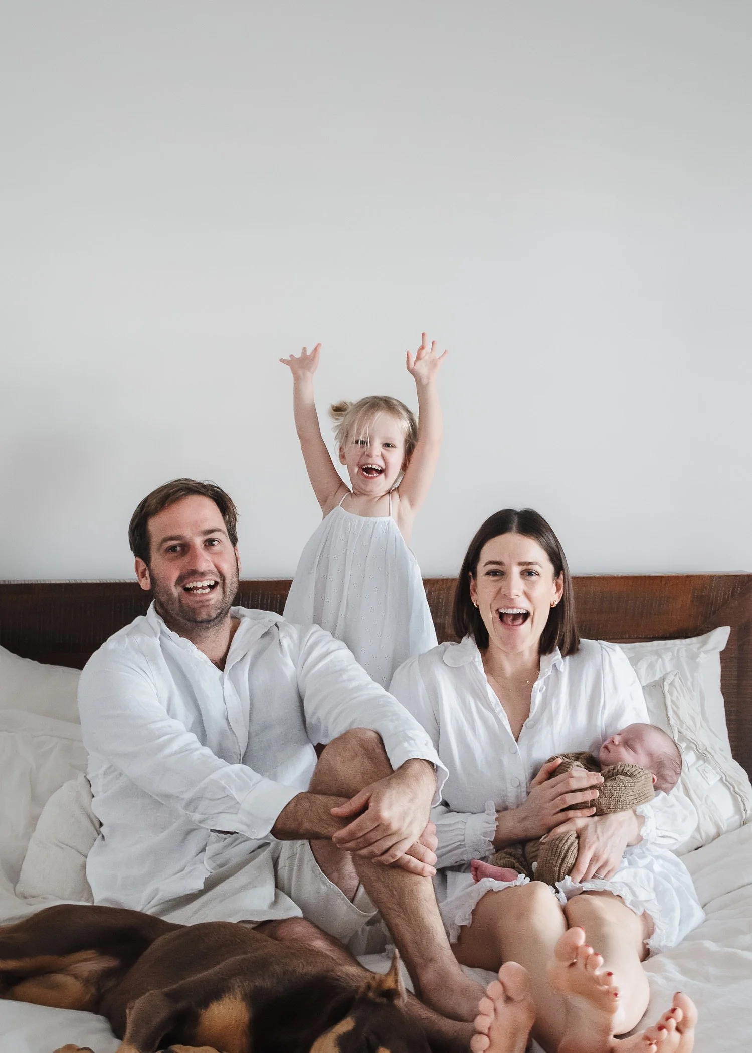 A happy family consisting of a father, mother, and two young children, sitting on a bed in a bedroom. The father has short dark hair, the mother has shoulder-length dark hair, and the children are a toddler girl with blonde hair and a newborn baby. T