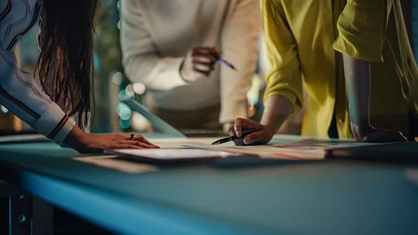 Three people leaning over a table reviewing documents with pens during a meeting.