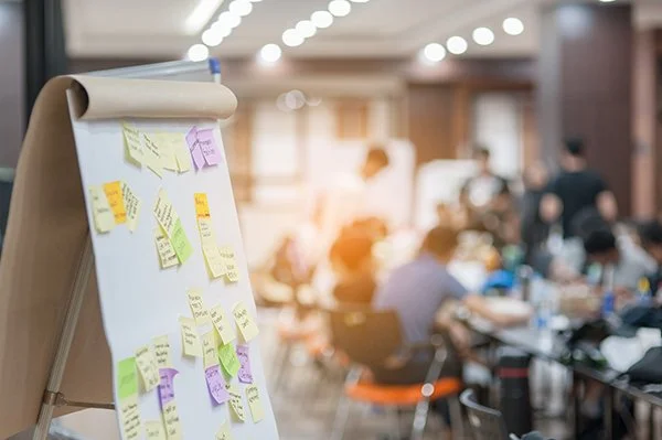 Flip chart with colorful sticky notes in focus with blurred people working in background.