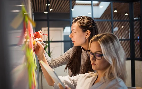 Two women collaborating with sticky notes on a board in a modern office space.