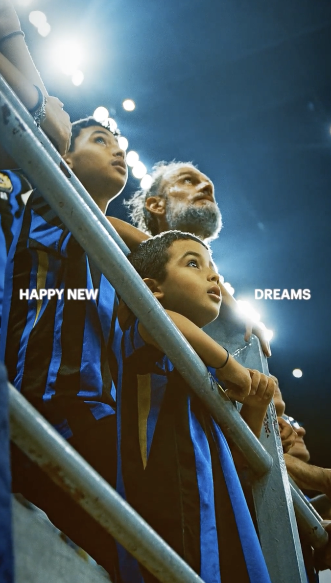 Tre spettatori, due adolescenti e un uomo più anziano, guardano verso il basso in un'arena sportiva sotto luci intense, con una scritta 'HAPPY NEW DREAMS' sovrapposta all'immagine.