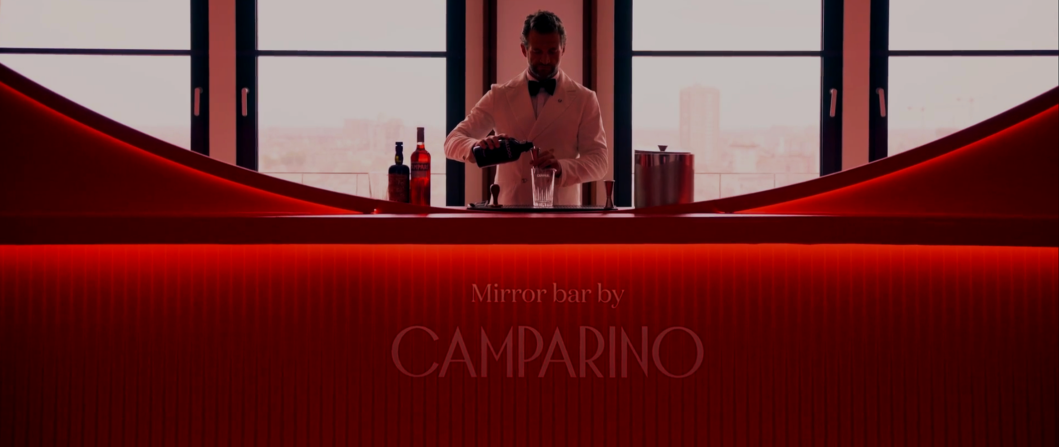 Barista in smokey white tuxedo preparing a cocktail at a modern bar with red lighting and large windows.