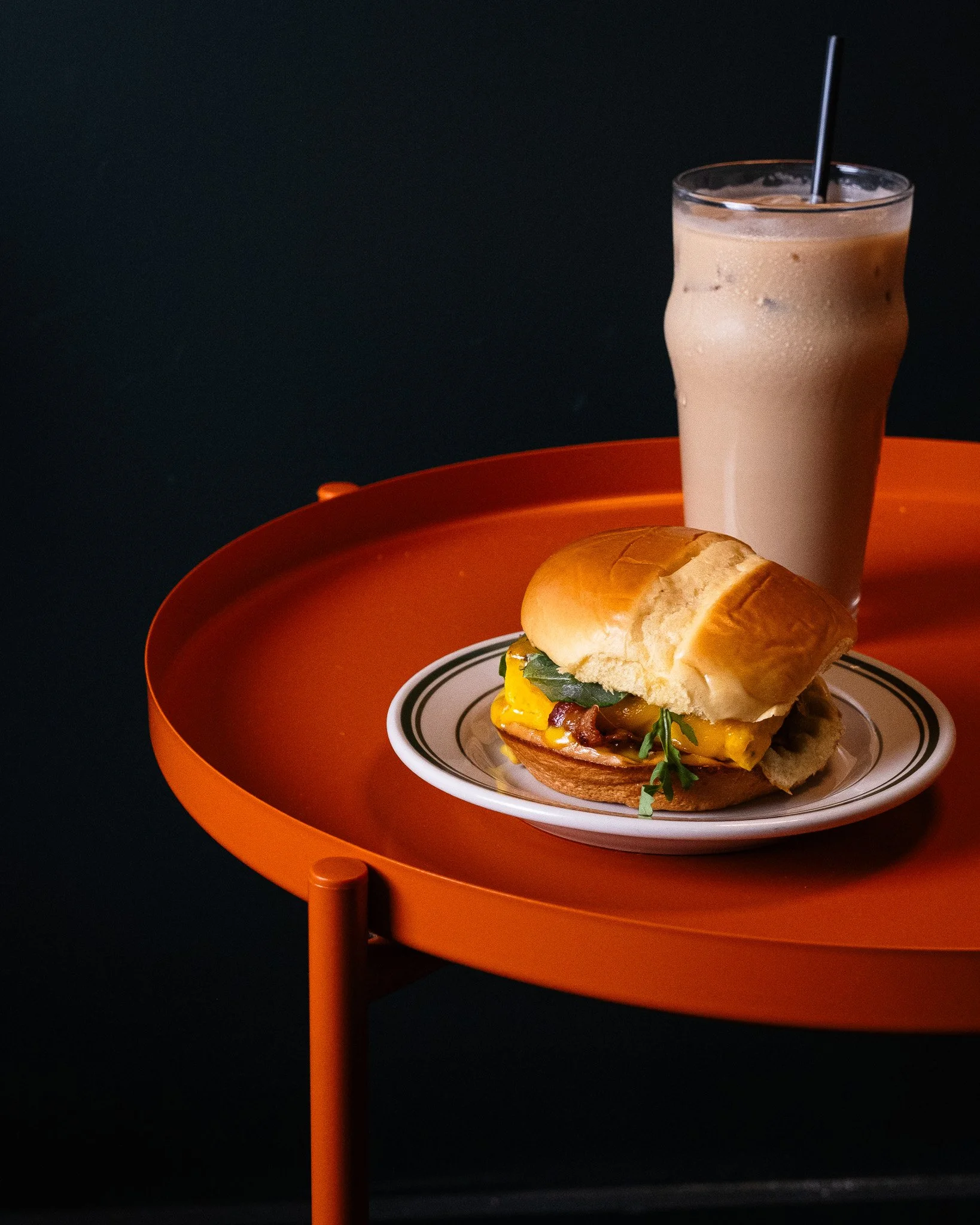 food photography for content day in Greenville, SC showing A sandwich with cheese and greens on a plate, and a glass of iced coffee with a straw on a round orange table.
