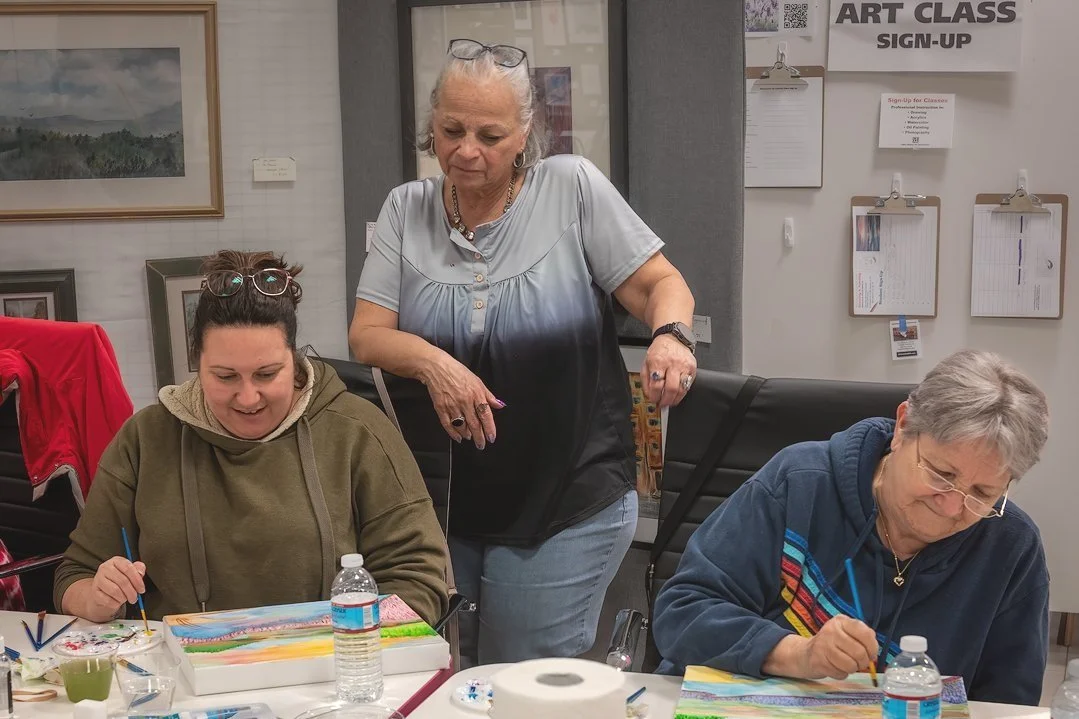 Three women painting at an art class, with framed artwork on the wall behind them.