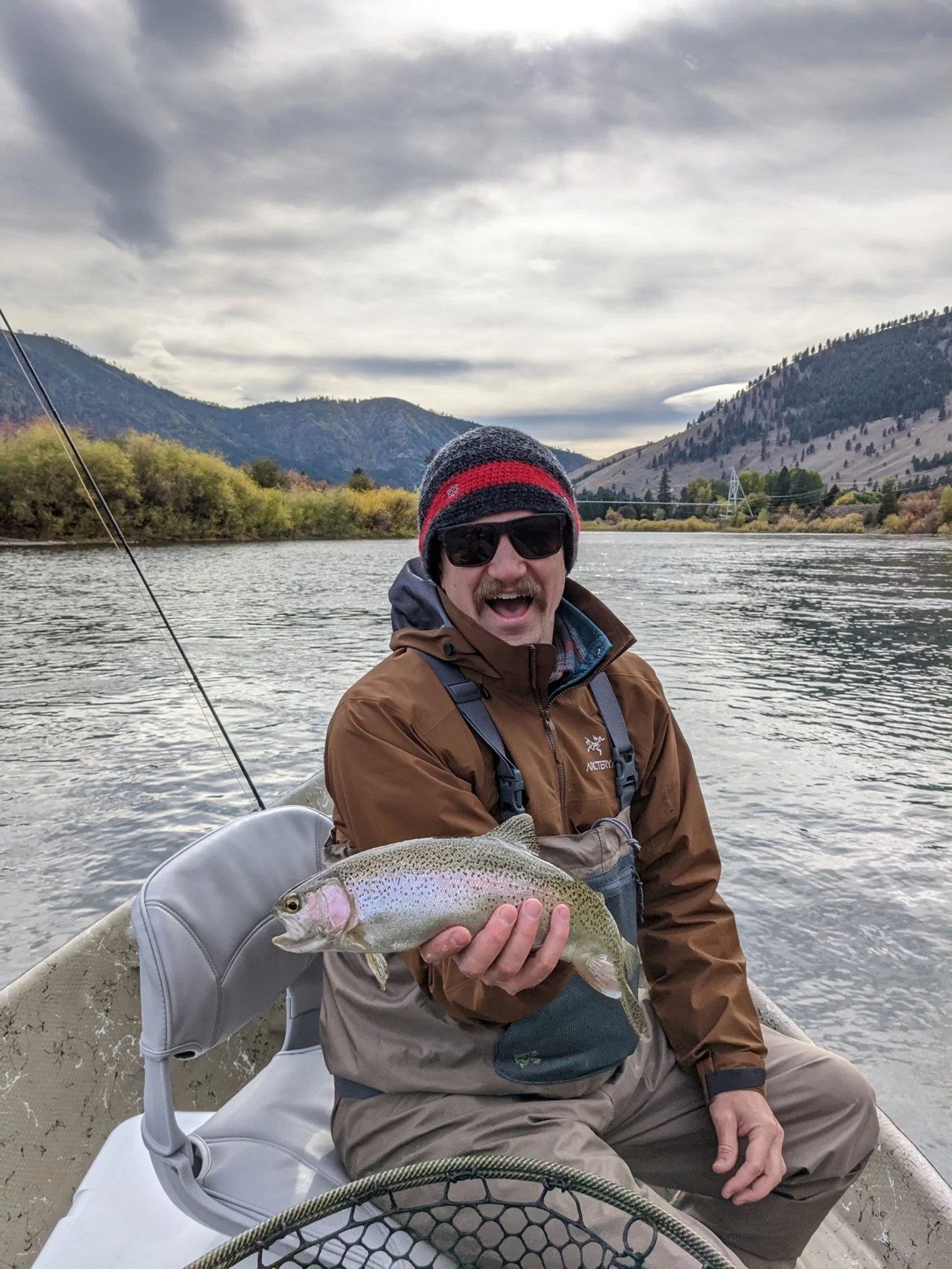 A man wearing sunglasses, a beanie, and a brown jacket holding a fish on a boat in a river with mountains and cloudy sky in the background.