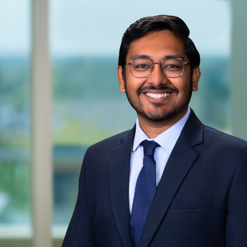 Professional man wearing glasses smiling in a business suit, office background with large windows.