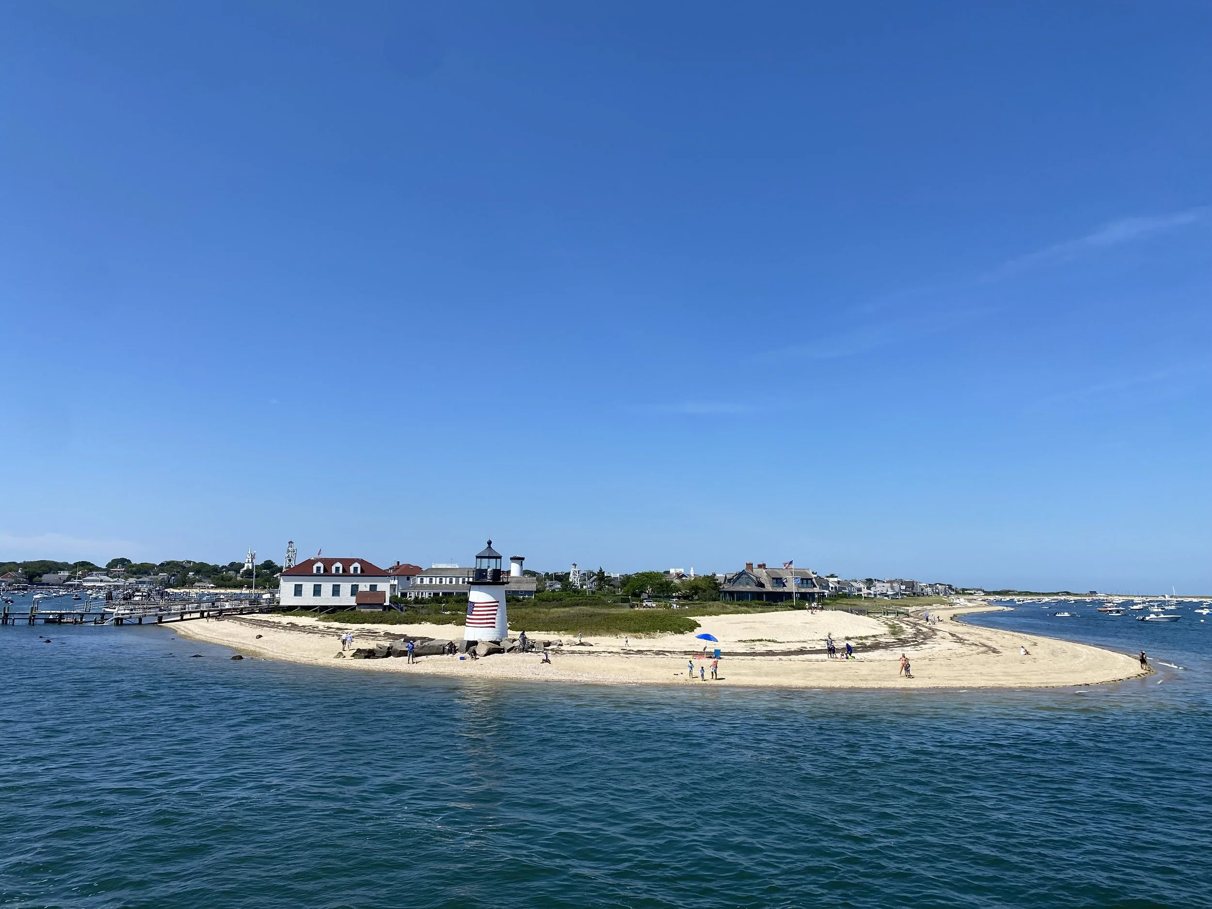 A small sandy beach with a lighthouse and several buildings in the background, with clear blue sky and calm ocean waters.