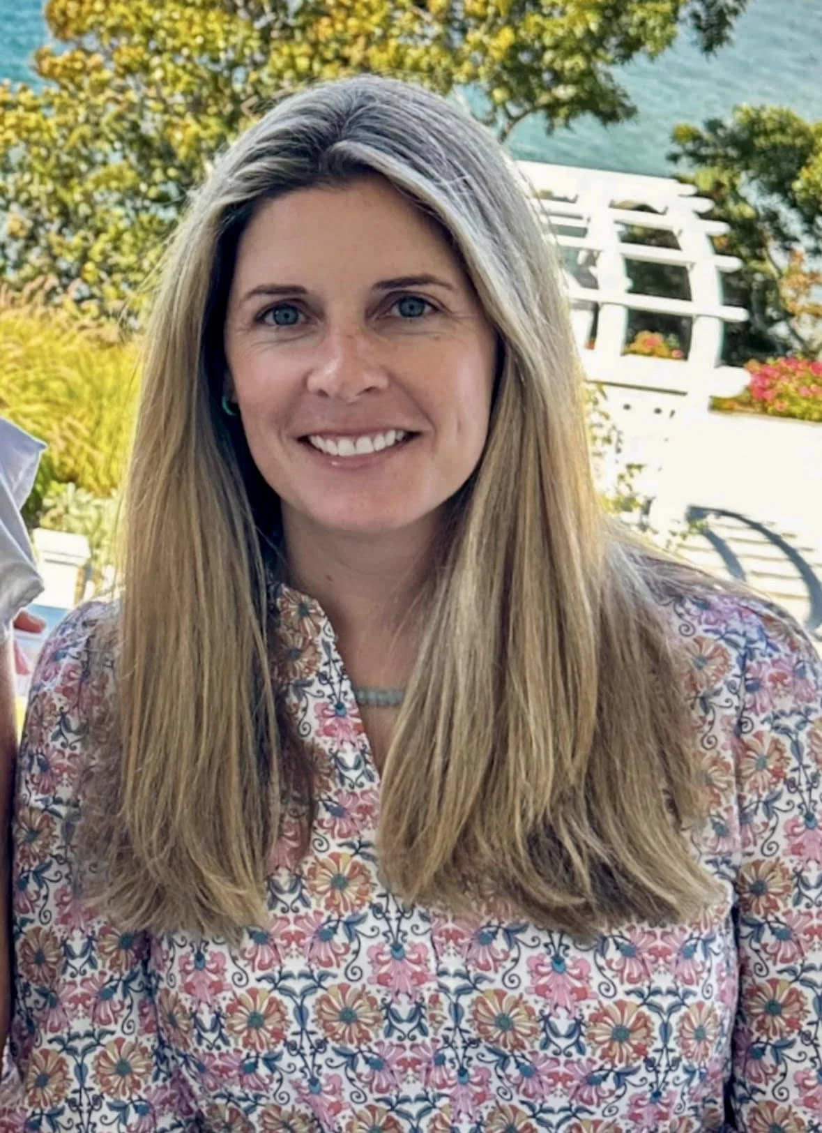 Woman with long hair wearing a floral shirt, standing outdoors with a blurred natural background.