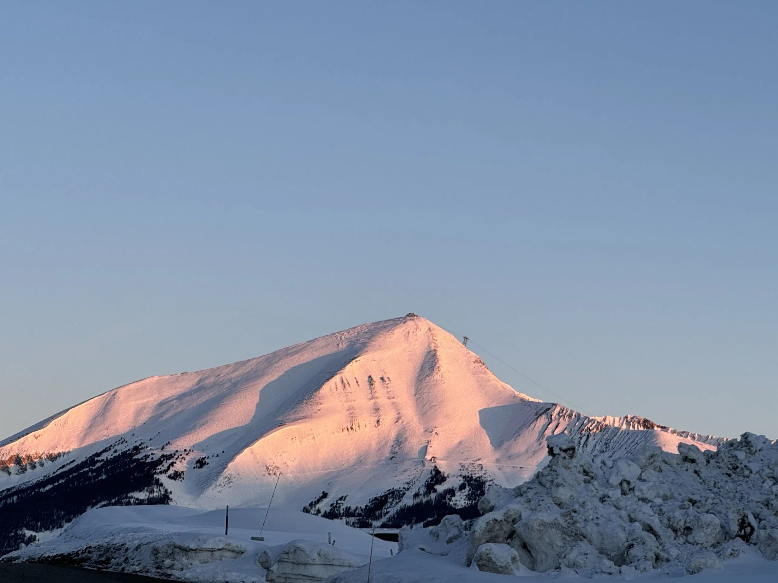 Snow-covered mountain peak with a clear sky at sunrise, casting a pinkish glow on the snow.