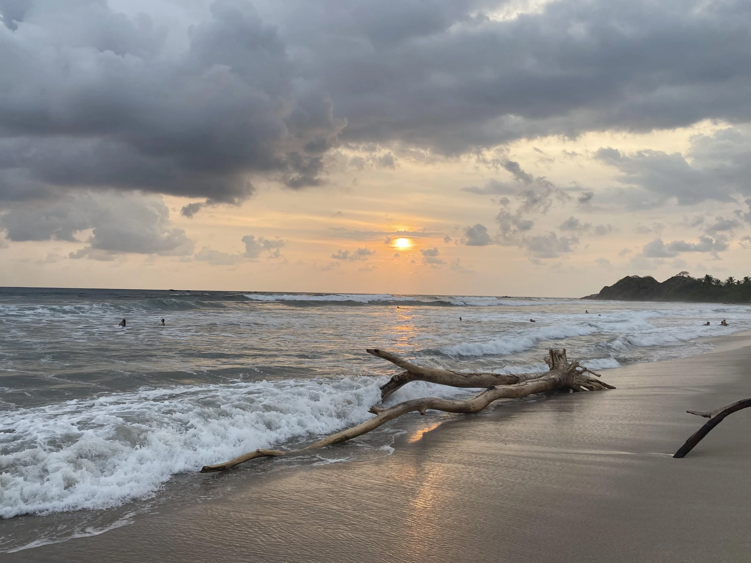 Sunset over an ocean with waves crashing on a sandy beach. Driftwood lies on the shore, and several people swim in the water. The sky is cloudy with a hint of sunlight.