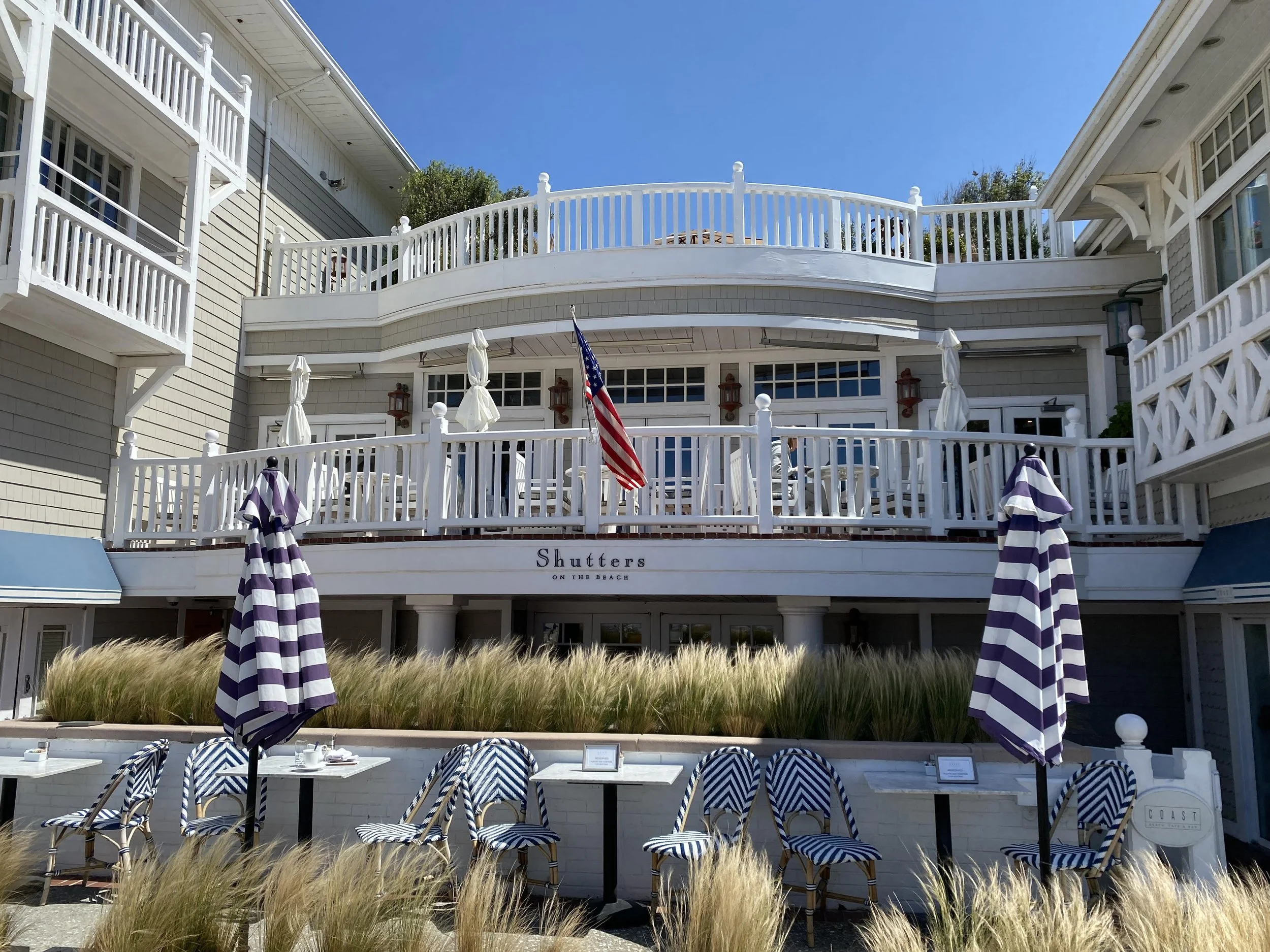Exterior of "Shutters on the Beach" hotel with American flag, white railings, striped umbrellas, and patio seating.