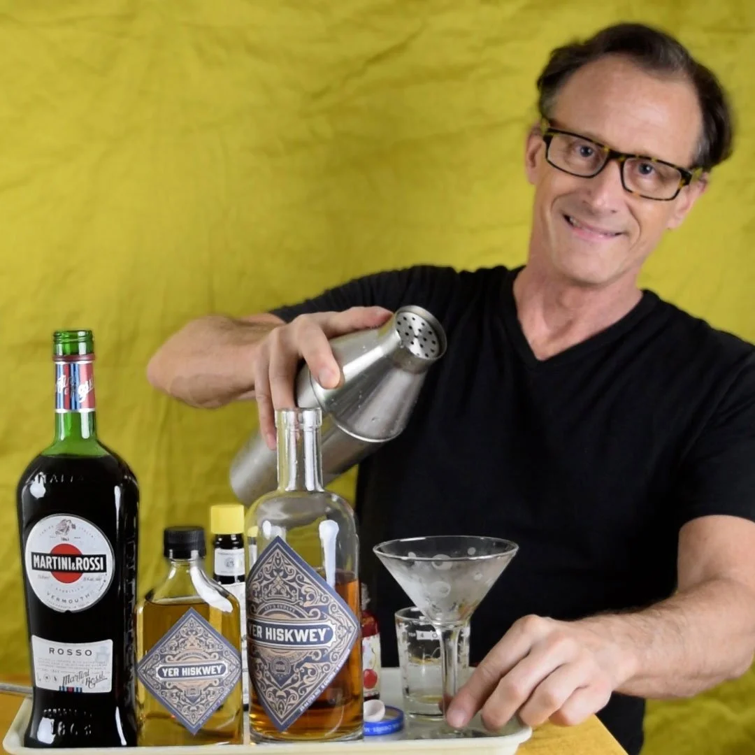 A man with glasses and a black shirt is pouring a clear liquid from a metal shaker into a martini glass. On the table, there are bottles of liquor, including Martini & Rossi vermouth and Yer Hiskey whiskey, along with some smaller bottles and a shot glass. The background is a yellow fabric or wall.