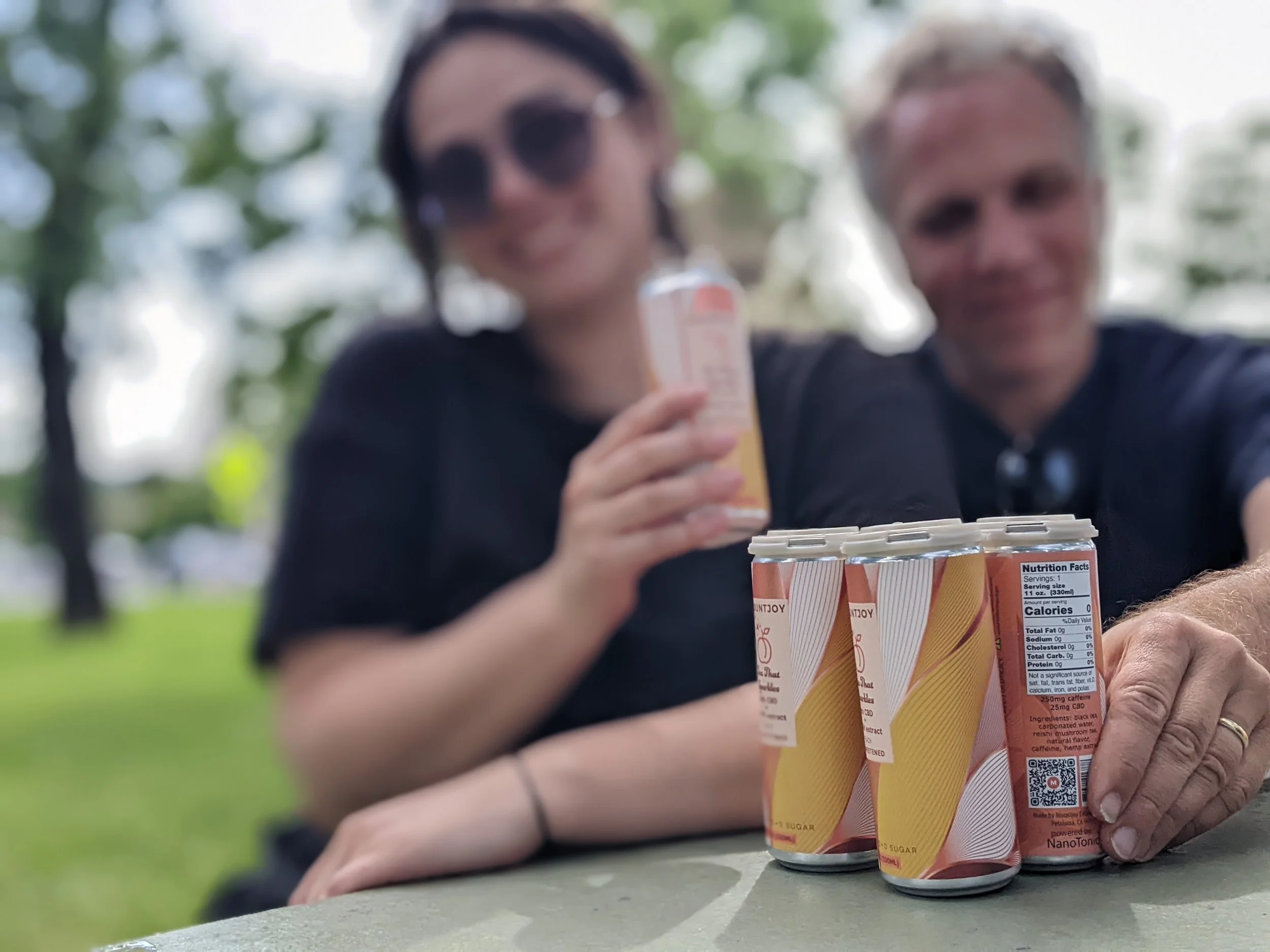 Two people sitting at a table outdoors, smiling, with a pyramid of beverage cans in the foreground. The background shows trees and a cloudy sky.