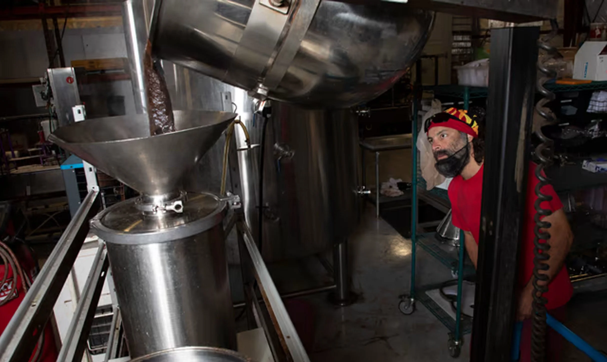 A man with a red bandana and red shirt peering around a metal industrial machine in a food production facility.