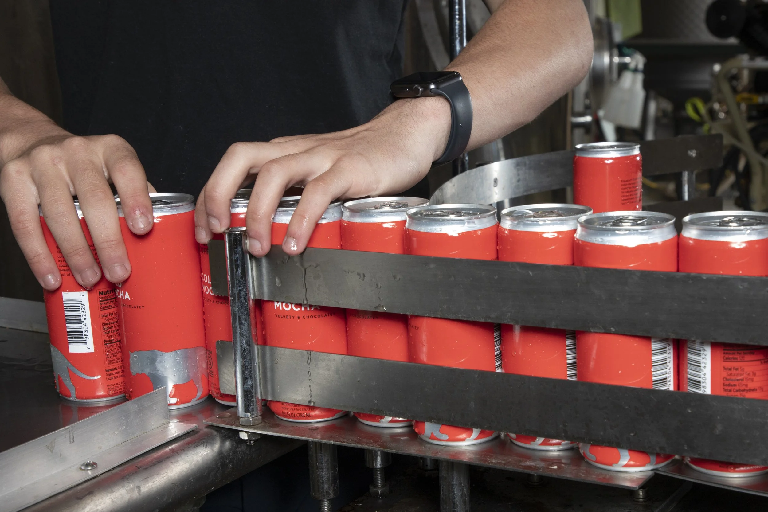 Person stacking cans of Mocha drink on a machine conveyor in a beverage production facility.