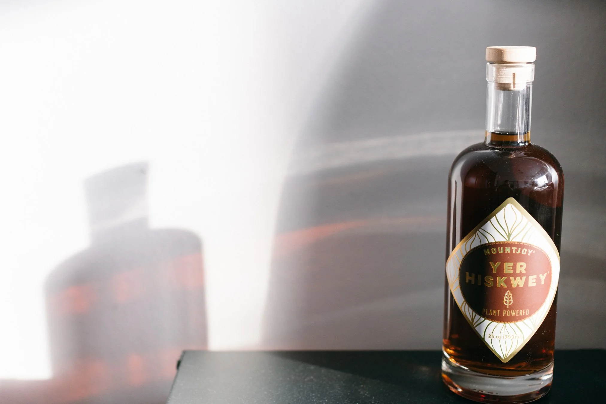 A bottle of Mountjoy Yer Hiskwey plant-powered beverage on a black surface with a shadow cast on a white wall.