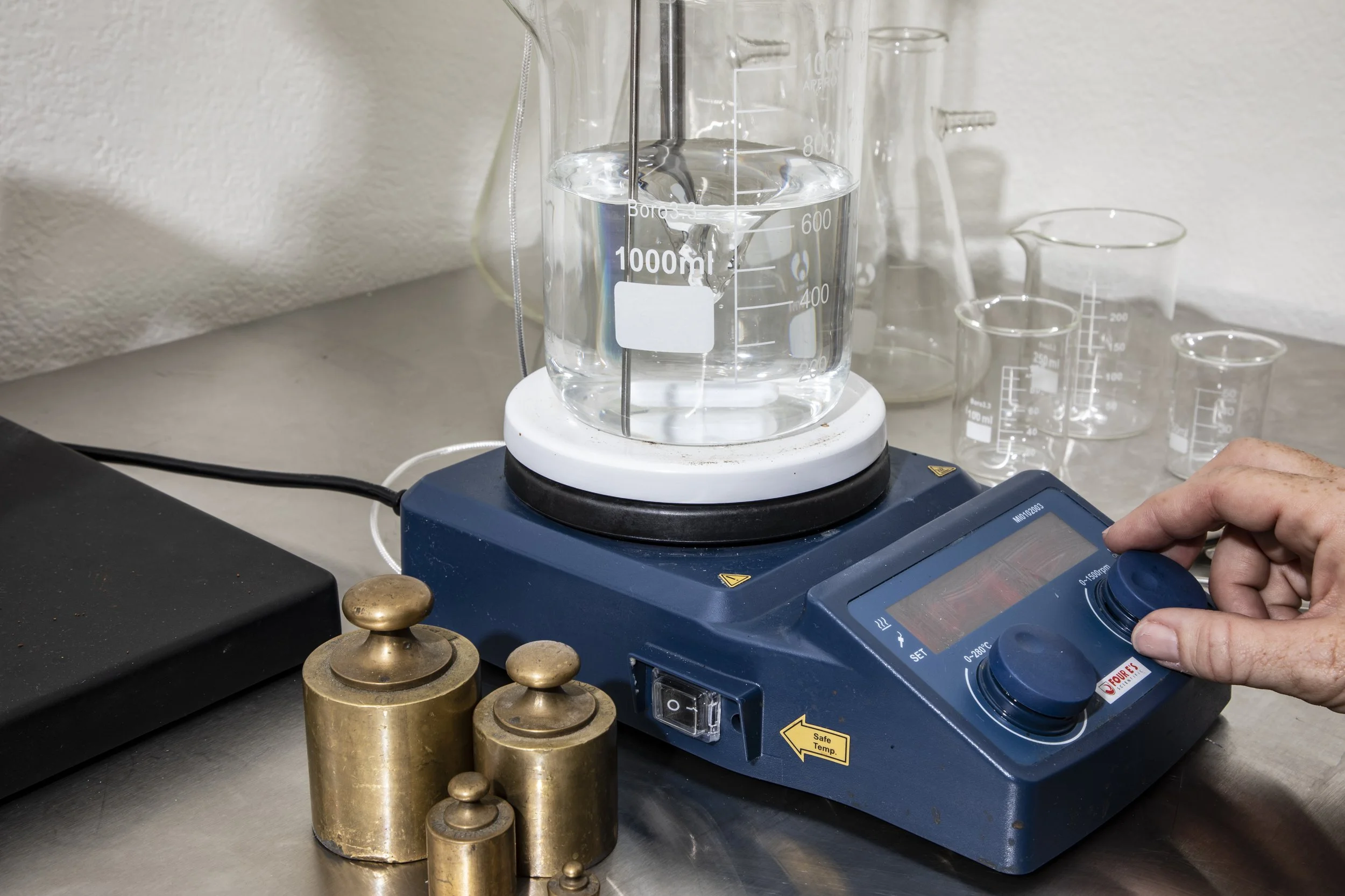 A laboratory setting with a digital hotplate stirrer, a beaker filled with a clear liquid, and various measuring cylinders and flasks on a gray countertop. A person is adjusting the controls on the hotplate.