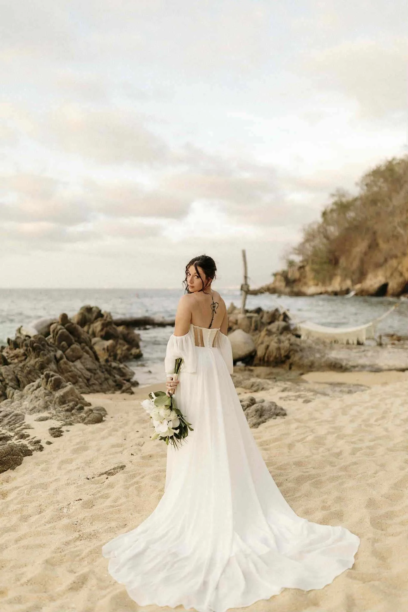 A bride in a white wedding dress holding a bouquet of white flowers on a sandy beach with rocks, ocean, and cloudy sky in the background.