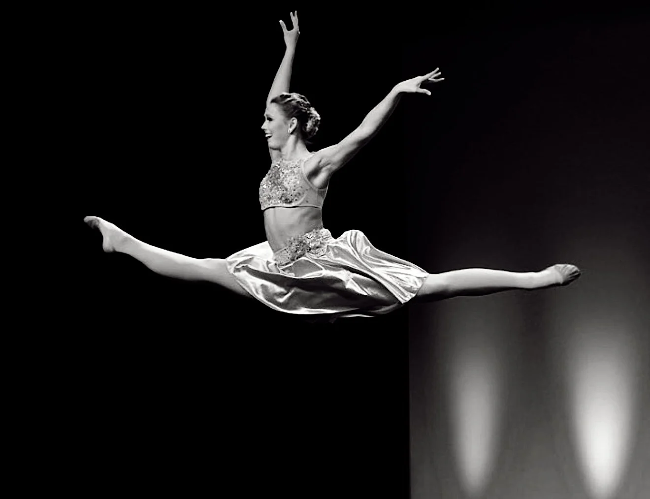 Black-and-white photo of a female ballet dancer mid-leap with arms extended upward and legs spread in a split, wearing a sequined top and flowing skirt against a dark background.