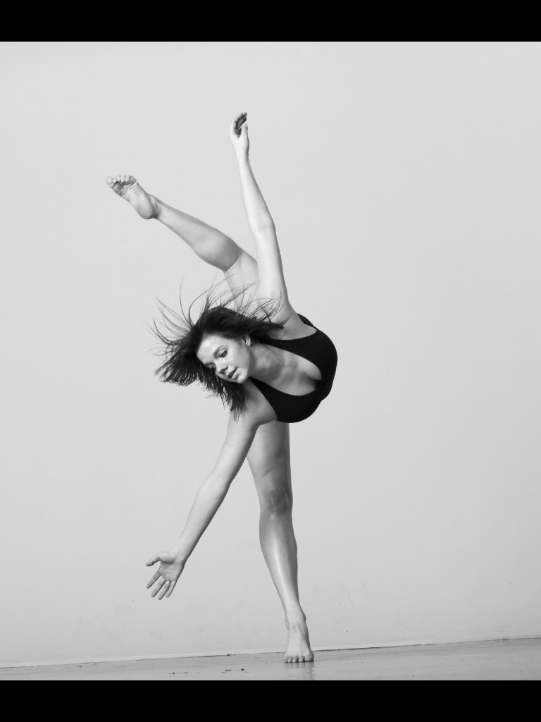 Black and white photo of a woman performing a dance pose, balancing on one foot with her other leg extended upward, and her arms extended outward.