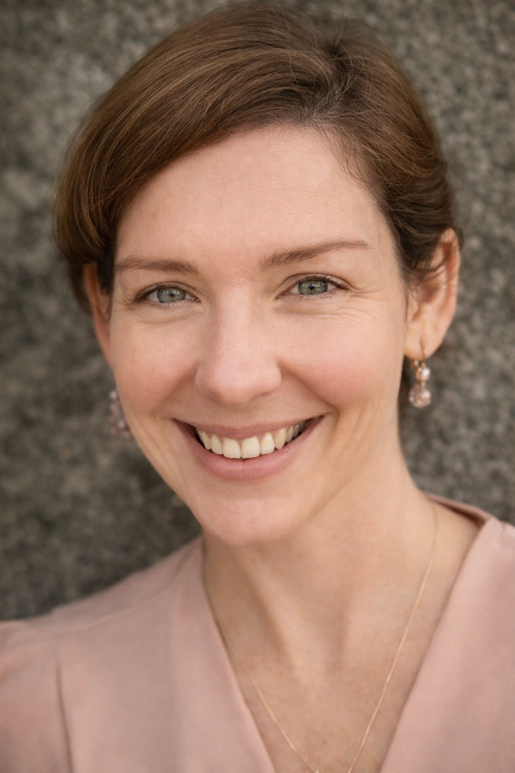 Close-up of a woman with short brown hair, blue eyes, smiling, wearing pearl earrings, a light pink top, and a thin gold necklace, standing against a stone background.