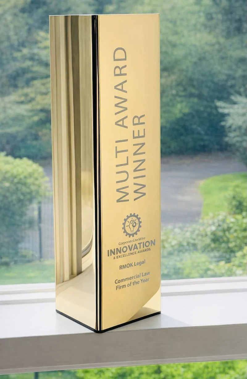 Tall gold-colored award plaque with the words 'MULTI AWARD WINNER' and below that, 'Corporate LiveWire INNOVATION & EXCELLENCE AWARDS Commercial Law Firm of the Year' in black text, placed on a windowsill with a blurred view of trees outside in the background.