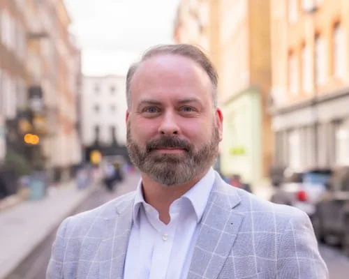 A man with a beard and short hair wearing a light-colored checkered blazer and white shirt, standing outdoors on a city street.