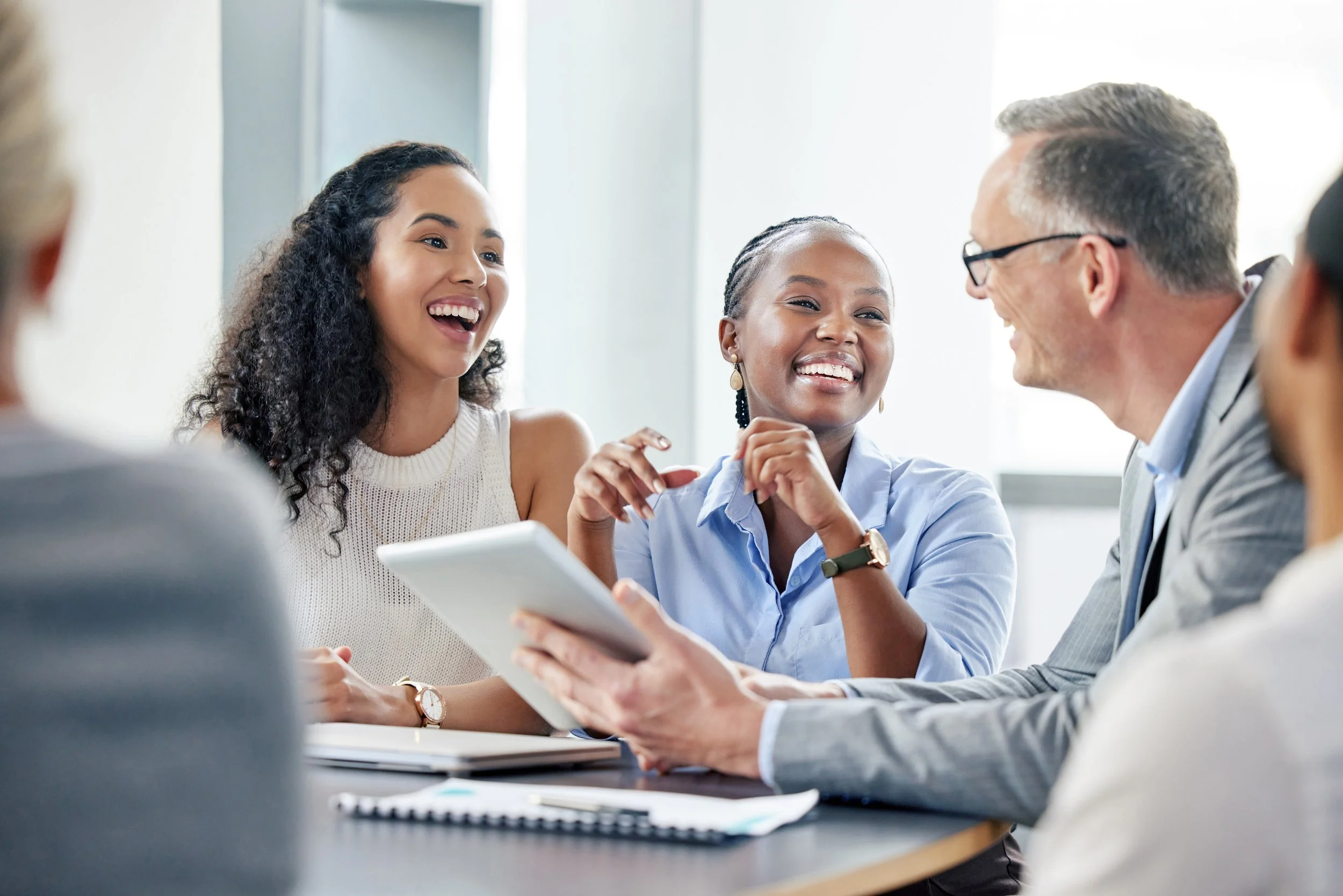 Business professionals in a meeting room engaged in a lively discussion, smiling and using a tablet and notebooks.