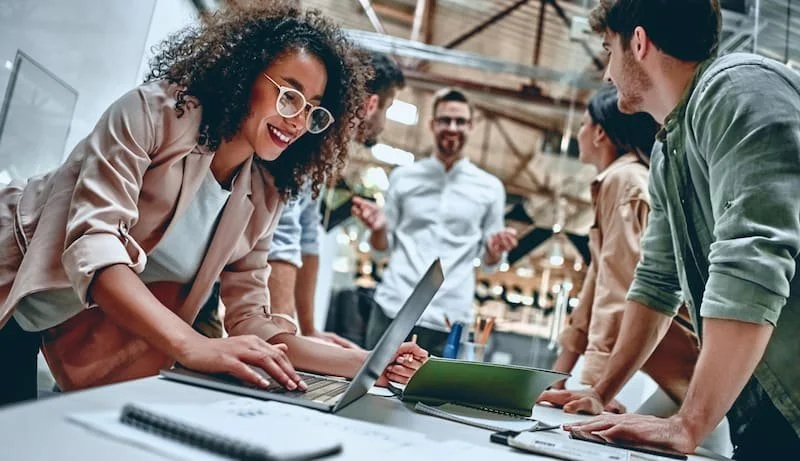 A group of young professionals gathered around a laptop in a modern office, engaging in a discussion and smiling.