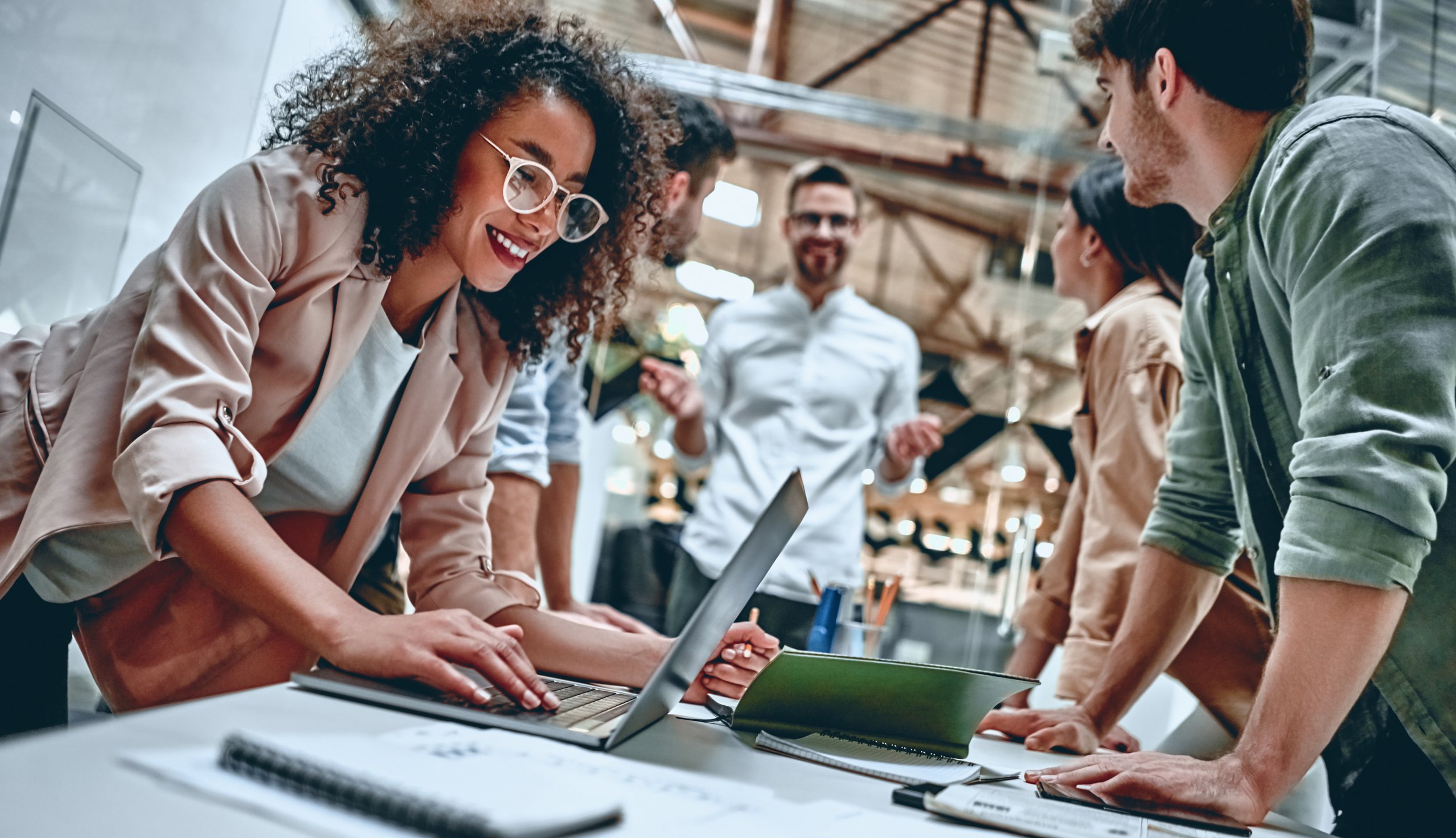 Group of diverse young professionals collaborating around a table with a laptop, notebooks, and pens in an industrial-style office.