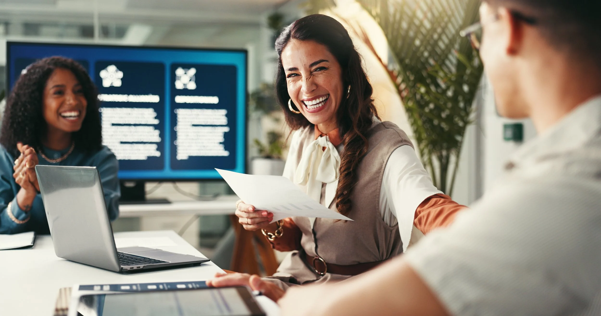 Three women in a professional office setting, with one woman in the foreground laughing and holding a paper, another woman in the background smiling, and a third person partially visible on the right, with a large screen displaying information behind them.