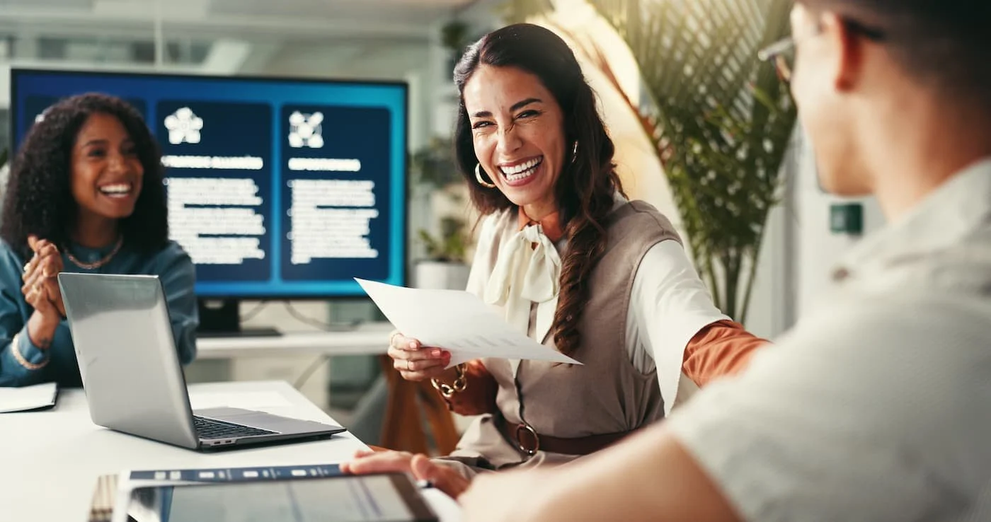Three women in a meeting, smiling and laughing, with laptops and documents on the table, in a bright office with plants and a large monitor in the background.