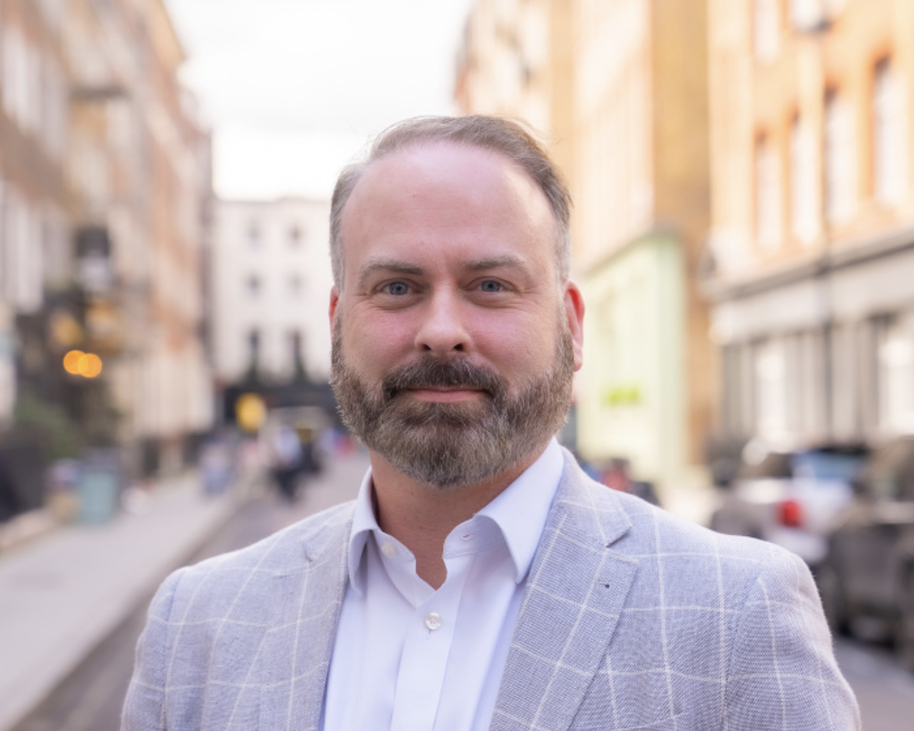 Rory O'Keeffe, Found and Principal Fractional General Counsel.  A man with a beard and short hair, wearing a light gray checkered blazer and white shirt, standing outdoors in an urban setting with blurred buildings and street in the background.