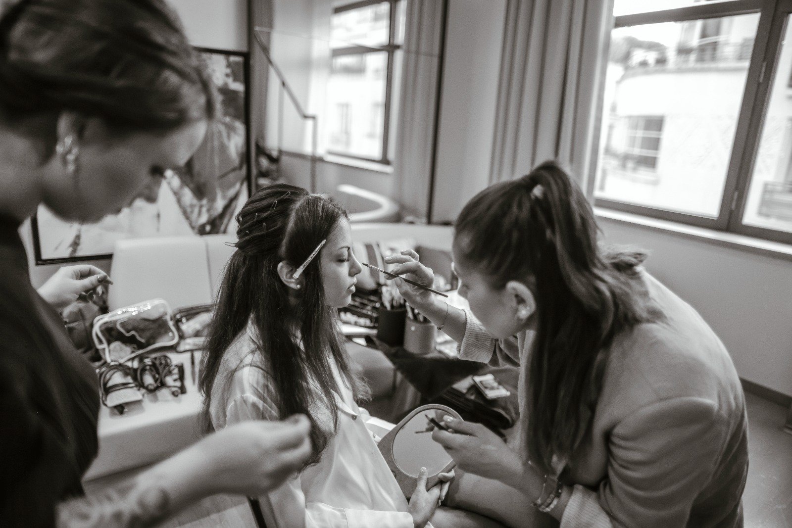 Une femme fait du maquillage à une autre femme assise, dans une pièce avec des fenêtres.elles sont près d'une table avec des kits de maquillage.