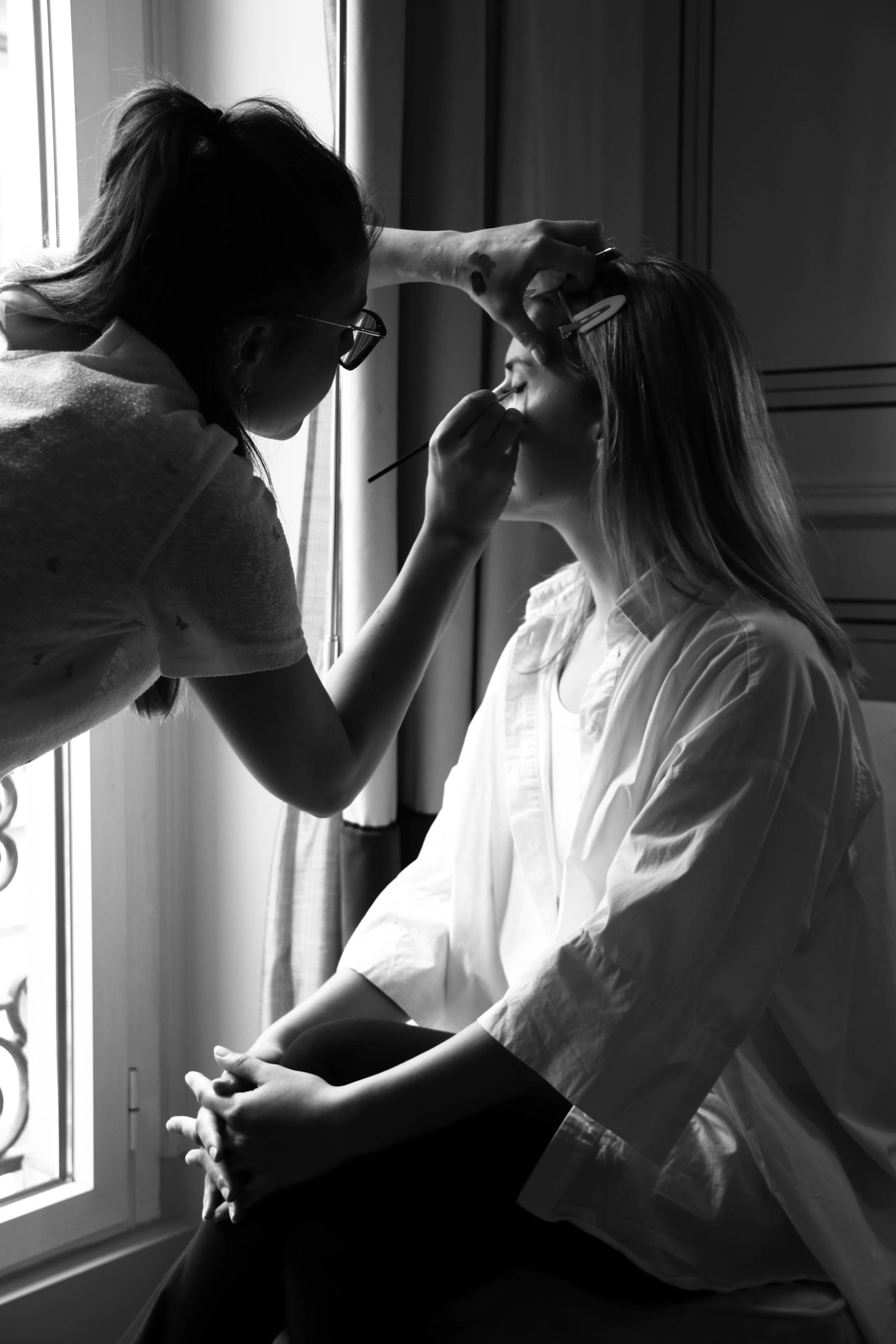 Une maquilleuse applique du maquillage à une femme assise près d'une fenêtre, en utilisant un pinceau, dans une pièce éclairée par la lumière naturelle.