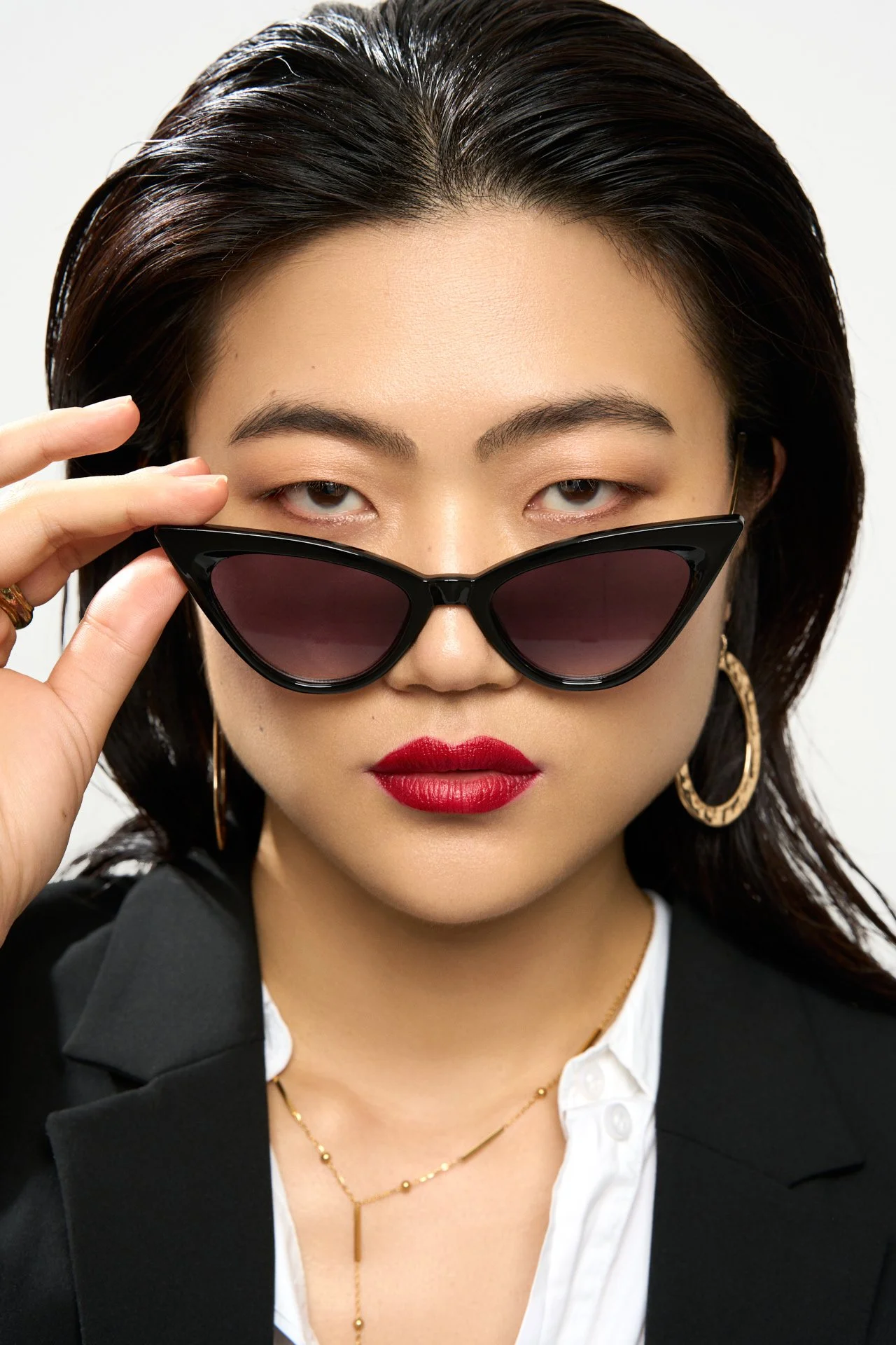 Une femme portant des lunettes de soleil noires, des boucles d'oreilles en or, un collier en or, avec un maquillage rouge à lèvres, regardant directement l'appareil.