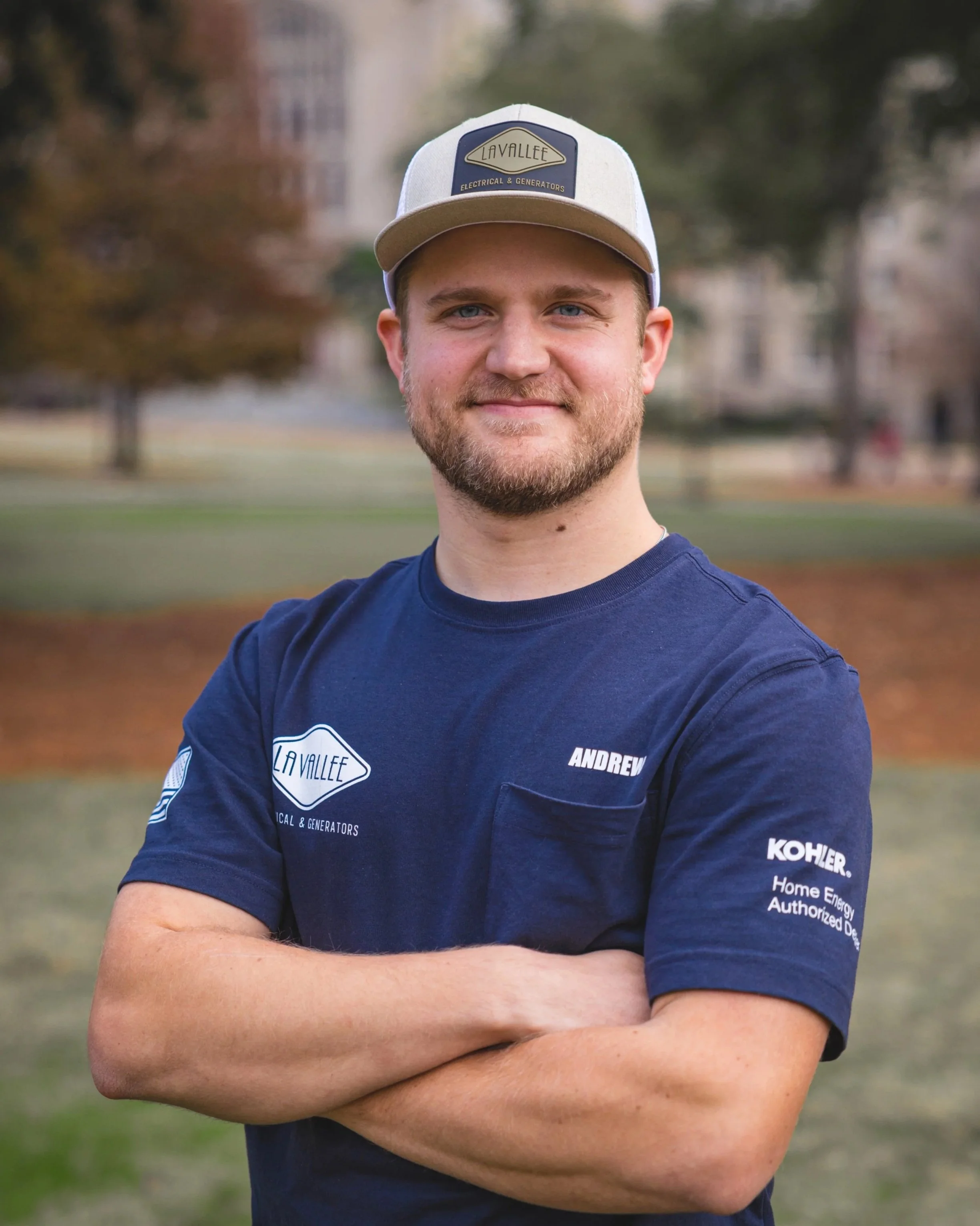 A young man with a beard and crossed arms standing outdoors in a park with trees and buildings in the background, wearing a navy blue t-shirt and a beige and gray cap with a logo.