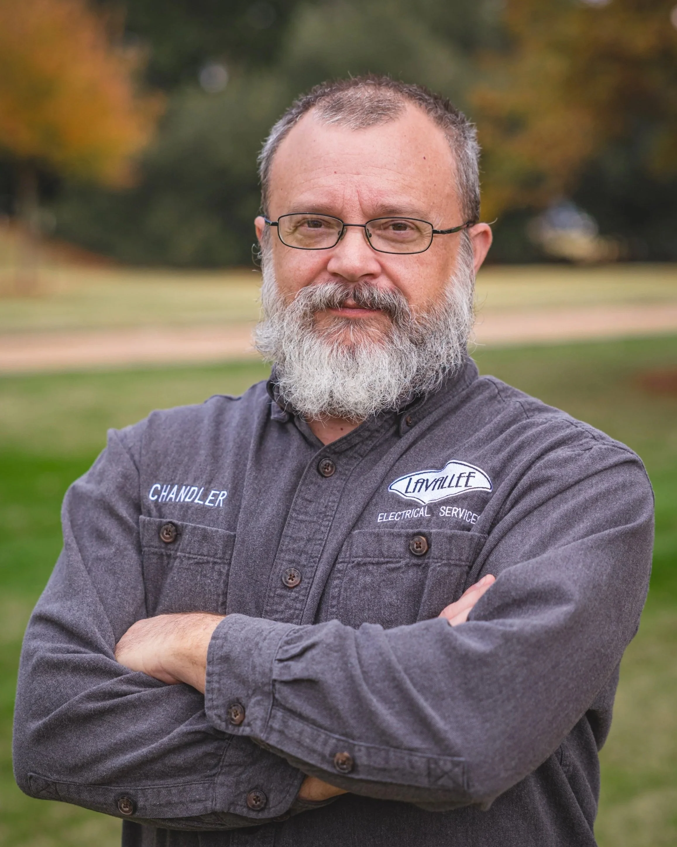 A middle-aged man with glasses and a full gray beard standing outdoors with his arms crossed, wearing a gray shirt with embroidered text 'Chandler' and 'LAVALLET Electrical Service' on it. The background shows a park with green grass and trees with autumn foliage.