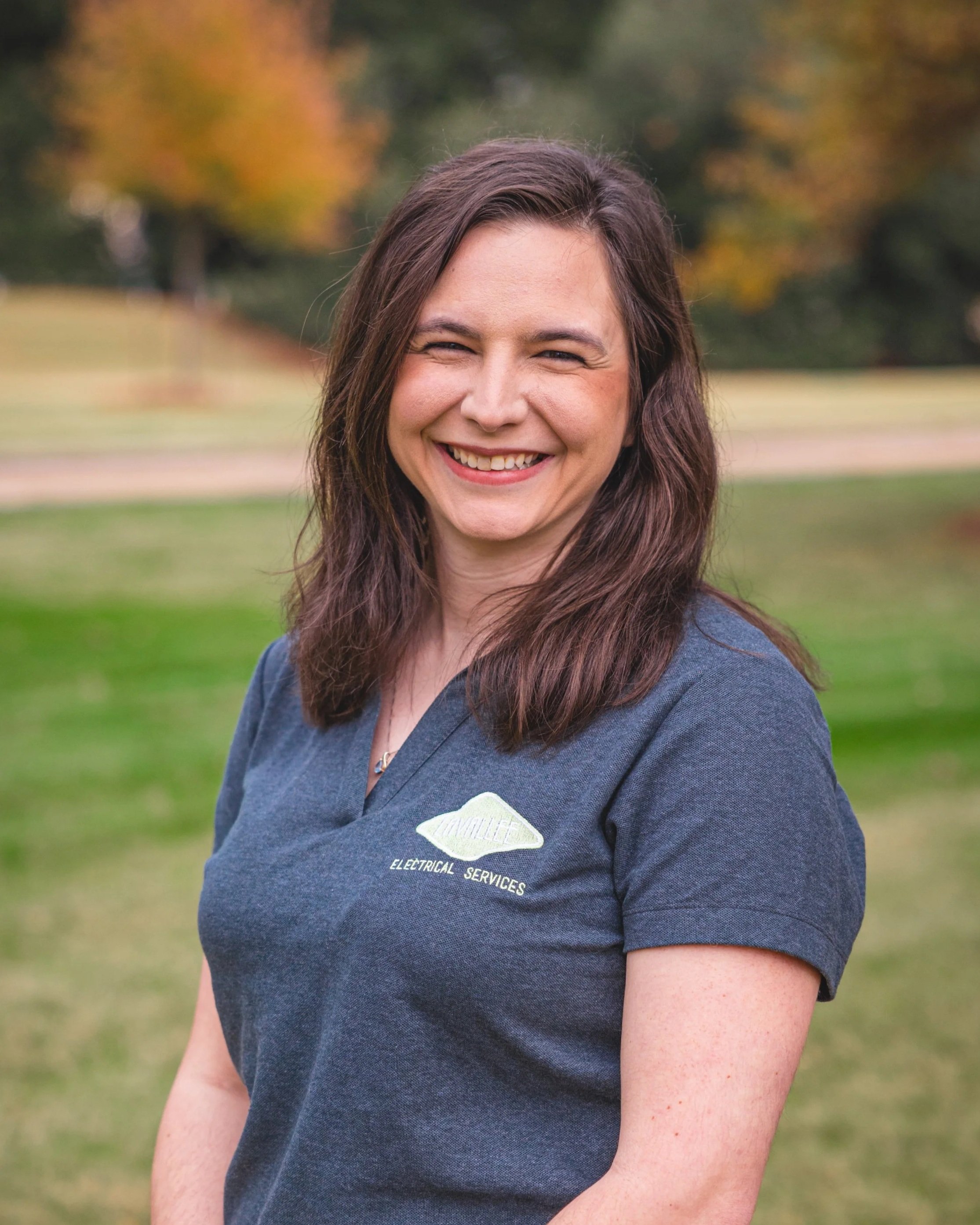 A smiling woman with brown hair, wearing a blue polo shirt with an embroidery of a light fixture and the words 'Electrical Services' on it, standing outdoors on a grassy area with trees in the background.