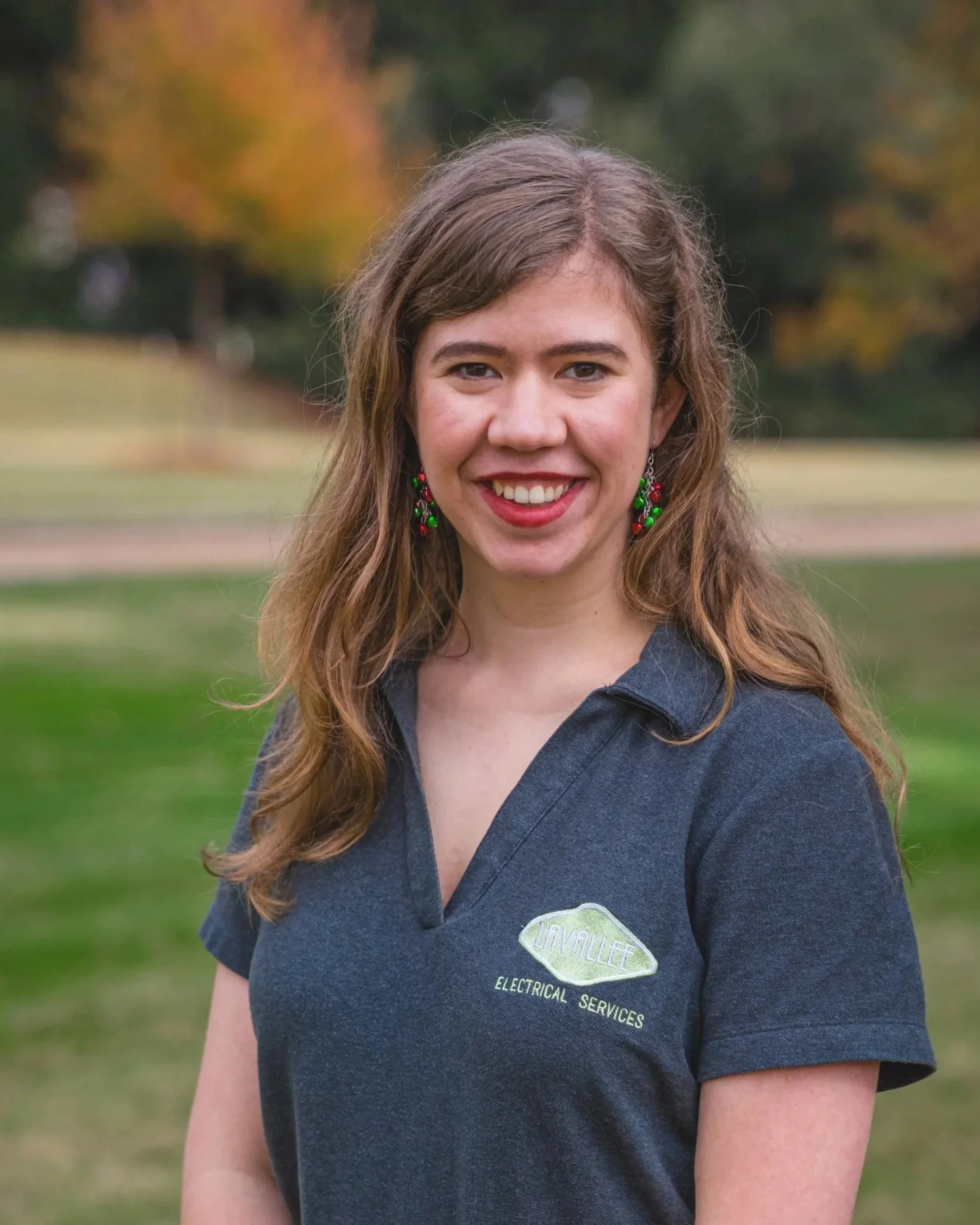 A young woman with long wavy brown hair smiling outdoors in a park. She is wearing red lipstick, green and red holiday earrings, and a dark gray polo shirt with a logo that reads 'LAVALLE ELECTRICAL SERVICES'.