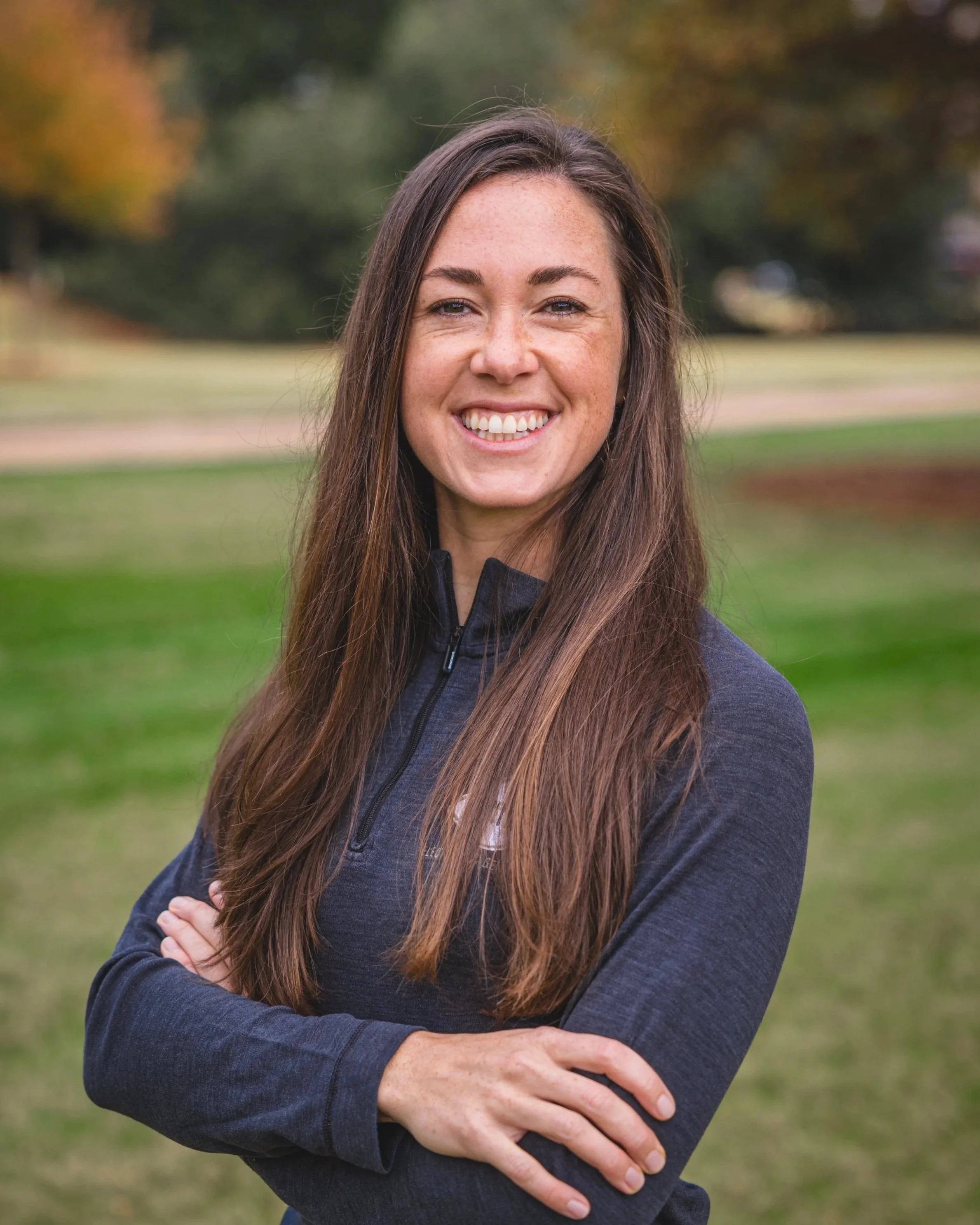A smiling woman with long brown hair, wearing a navy zip-up jacket, standing outdoors in a park with trees and grass in the background.