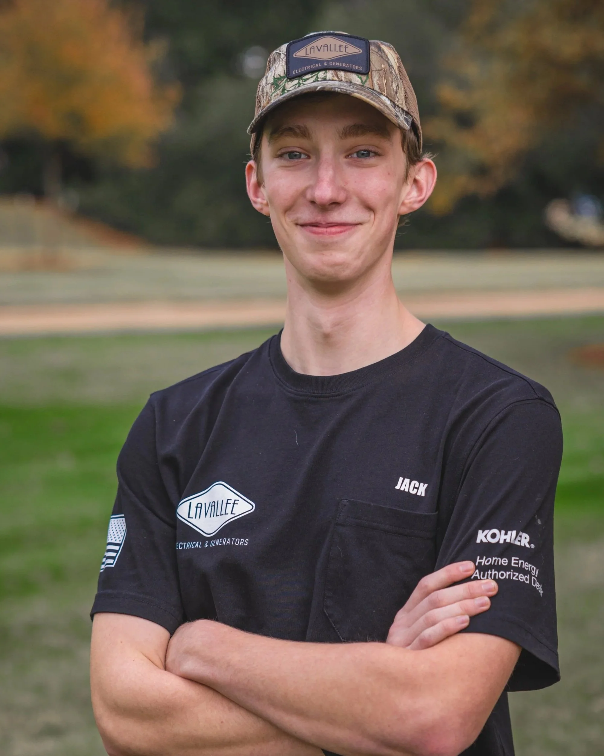 Young man standing outdoors with arms crossed, smiling, wearing a black shirt with sponsorship logos and a camouflage baseball cap, in a park with trees in the background.