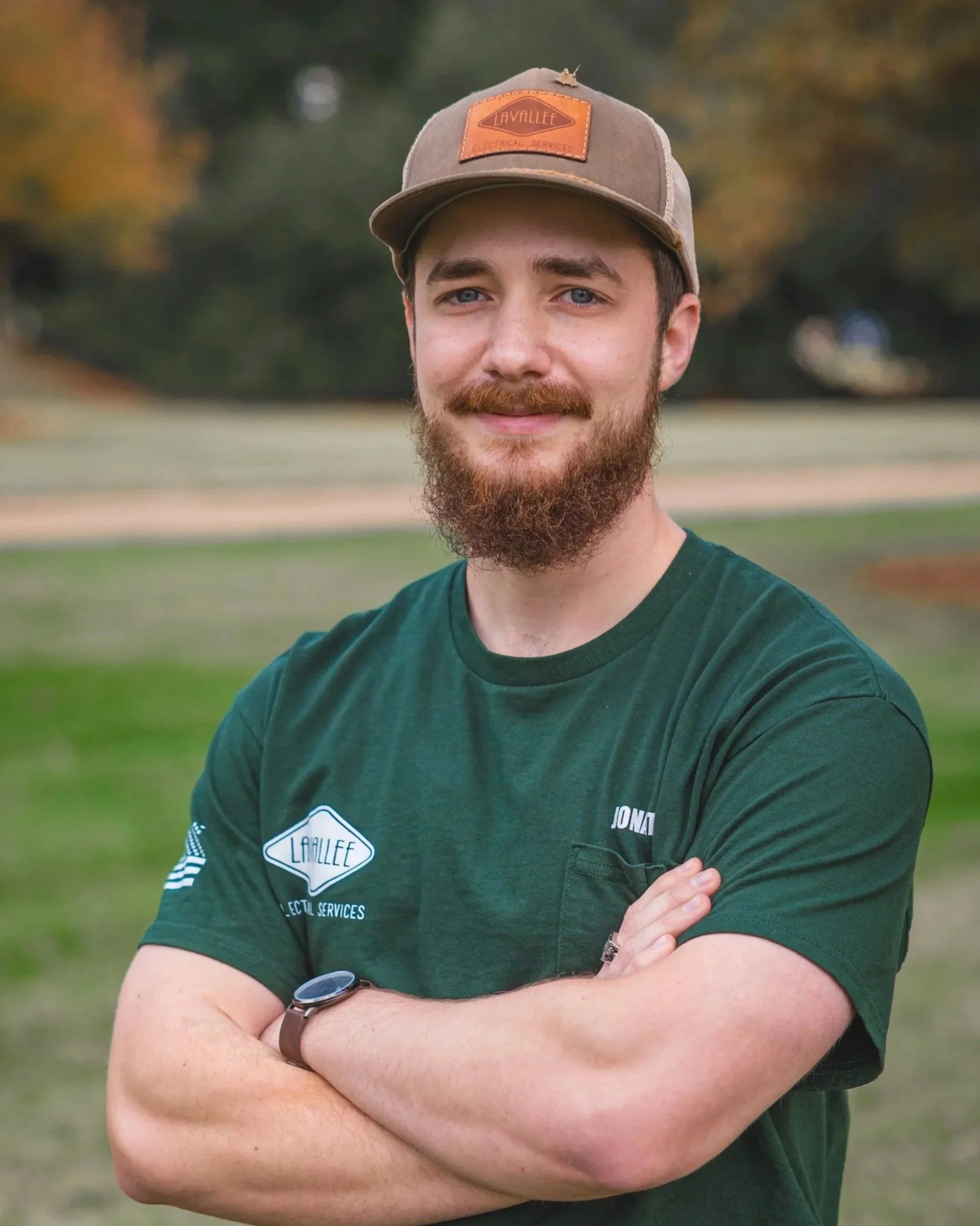 A man with a beard and mustache wearing a green T-shirt and a baseball cap, standing outdoors in a park with green grass and trees in the background.